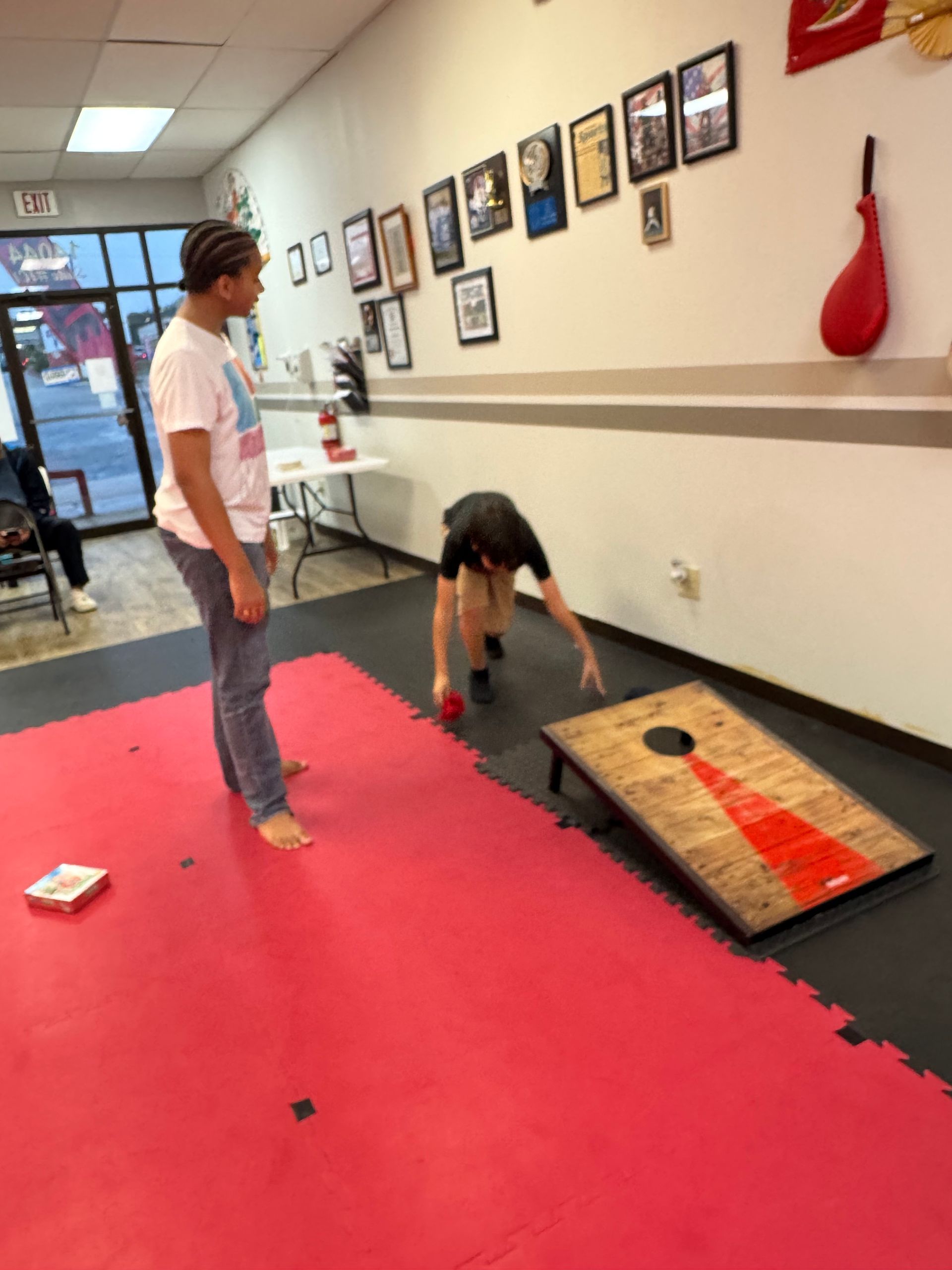 Two people in a gym hallway beside a red cornhole board, one standing and one bent over to throw.
