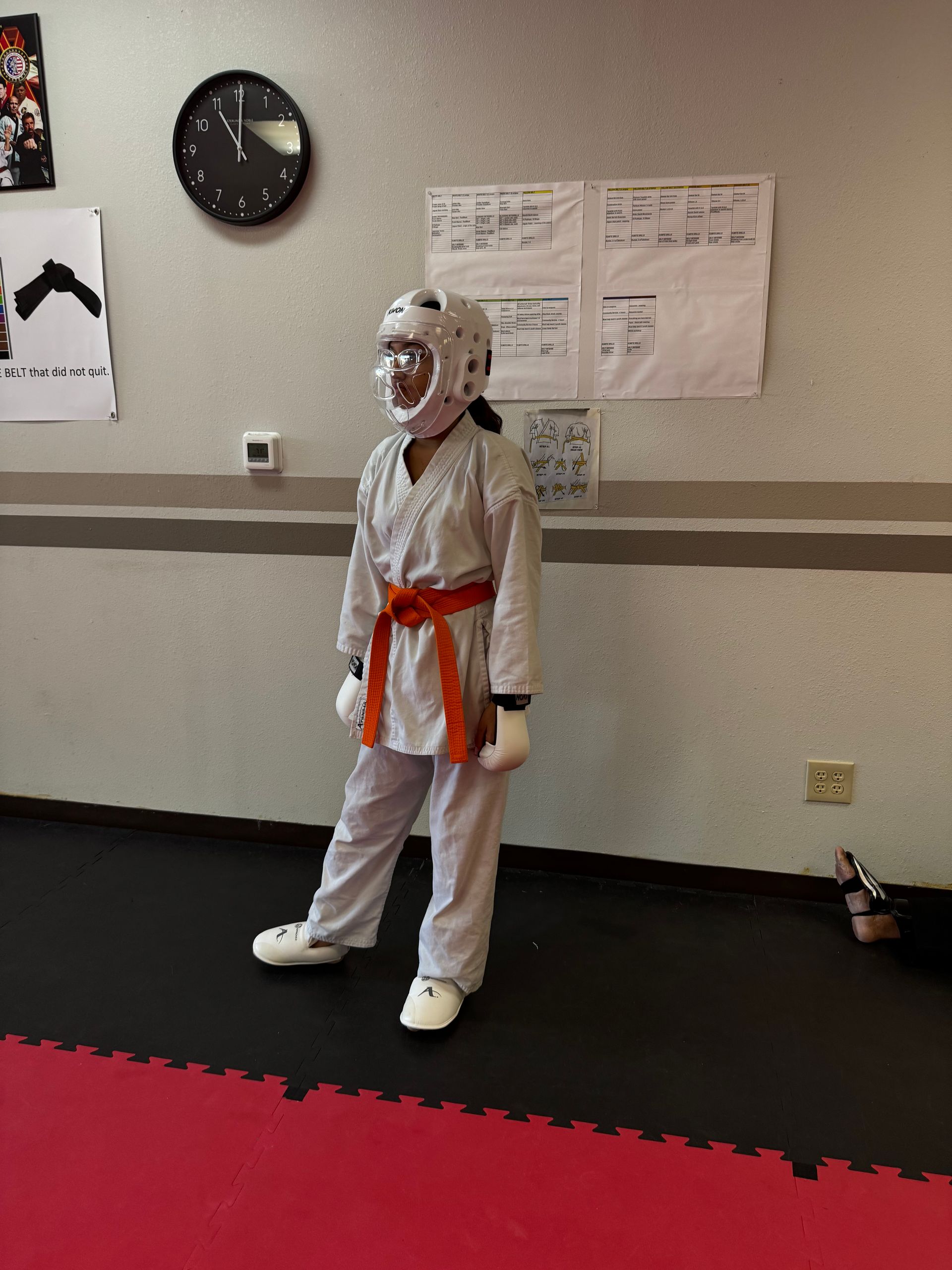 Child in white martial arts uniform with orange belt and protective headgear standing in a dojo.
