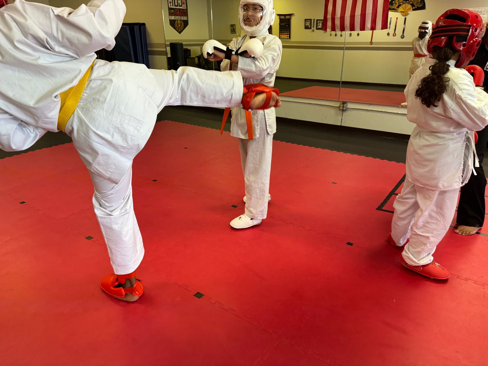Children in white karate uniforms spar on red mats in a dojo, one kicking while another wears red headgear.
