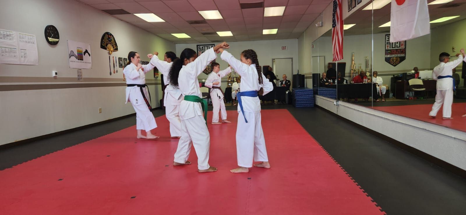 Martial arts students in white uniforms with colored belts practice in a mirrored dojo on a red mat.