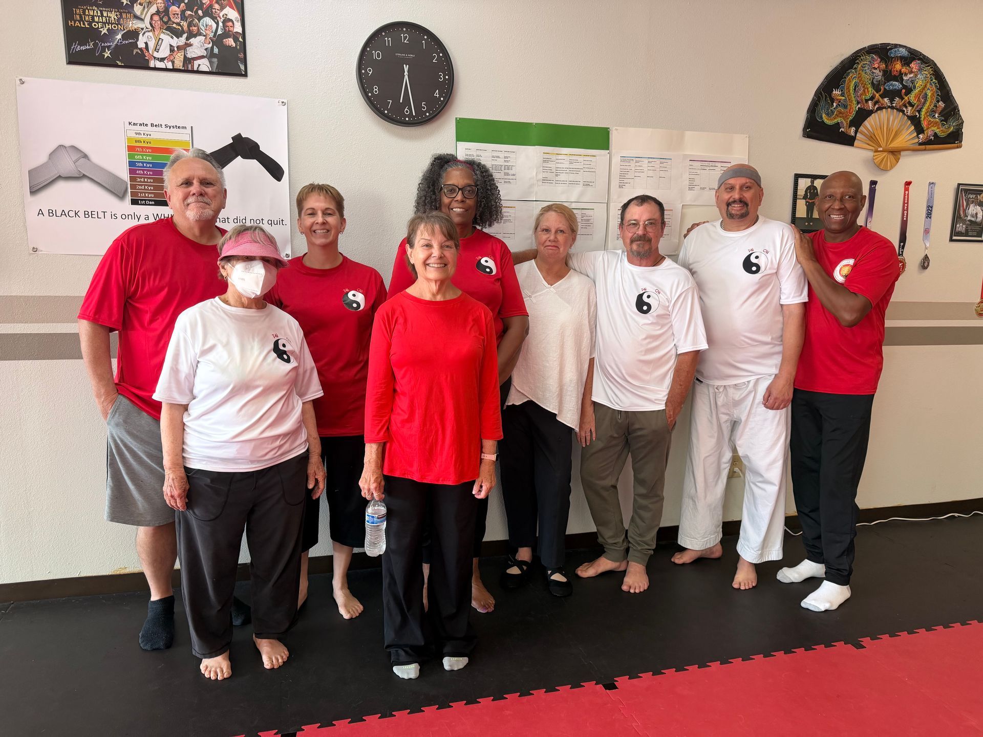 Group of people in red and white shirts posing in a martial arts studio with wall posters and clocks