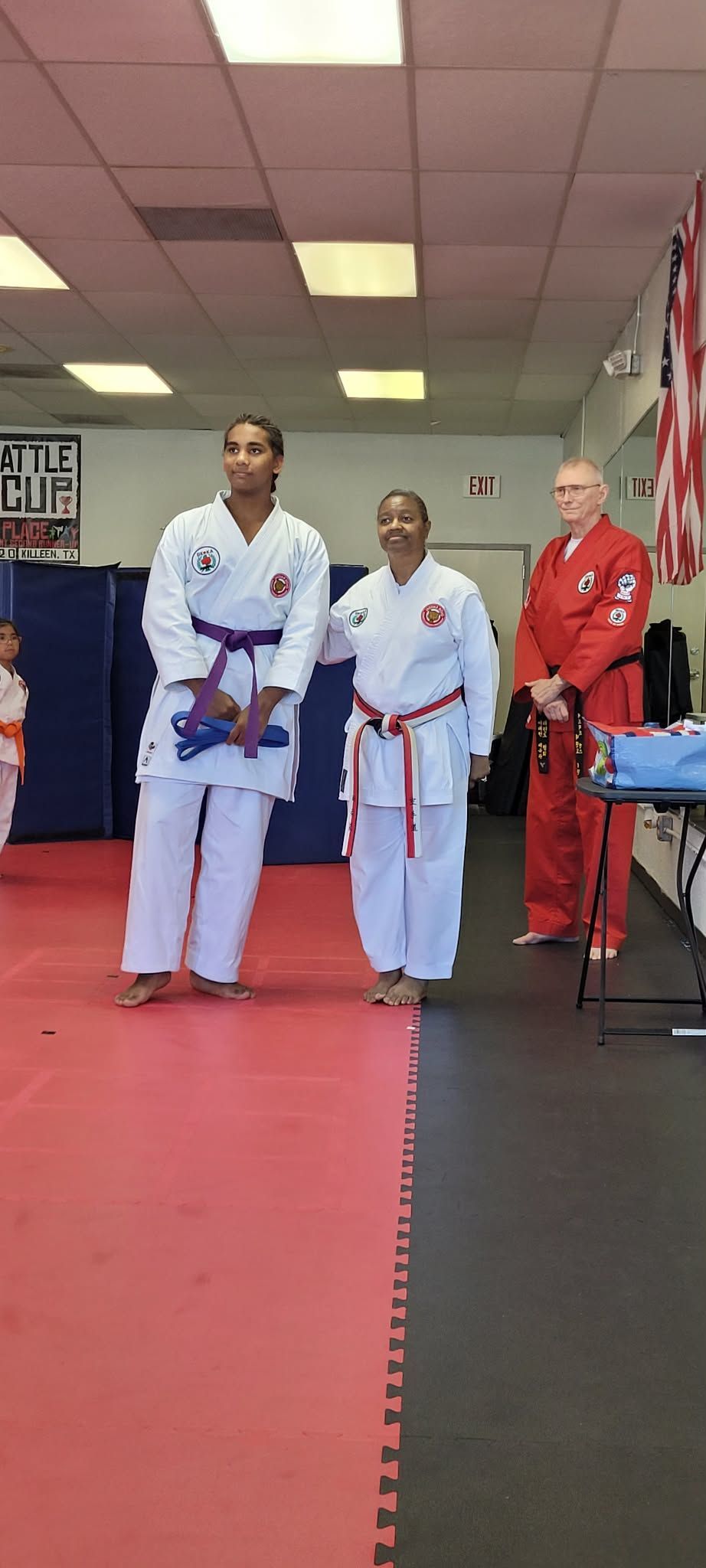 Three people in martial arts uniforms standing on a red mat in a dojo