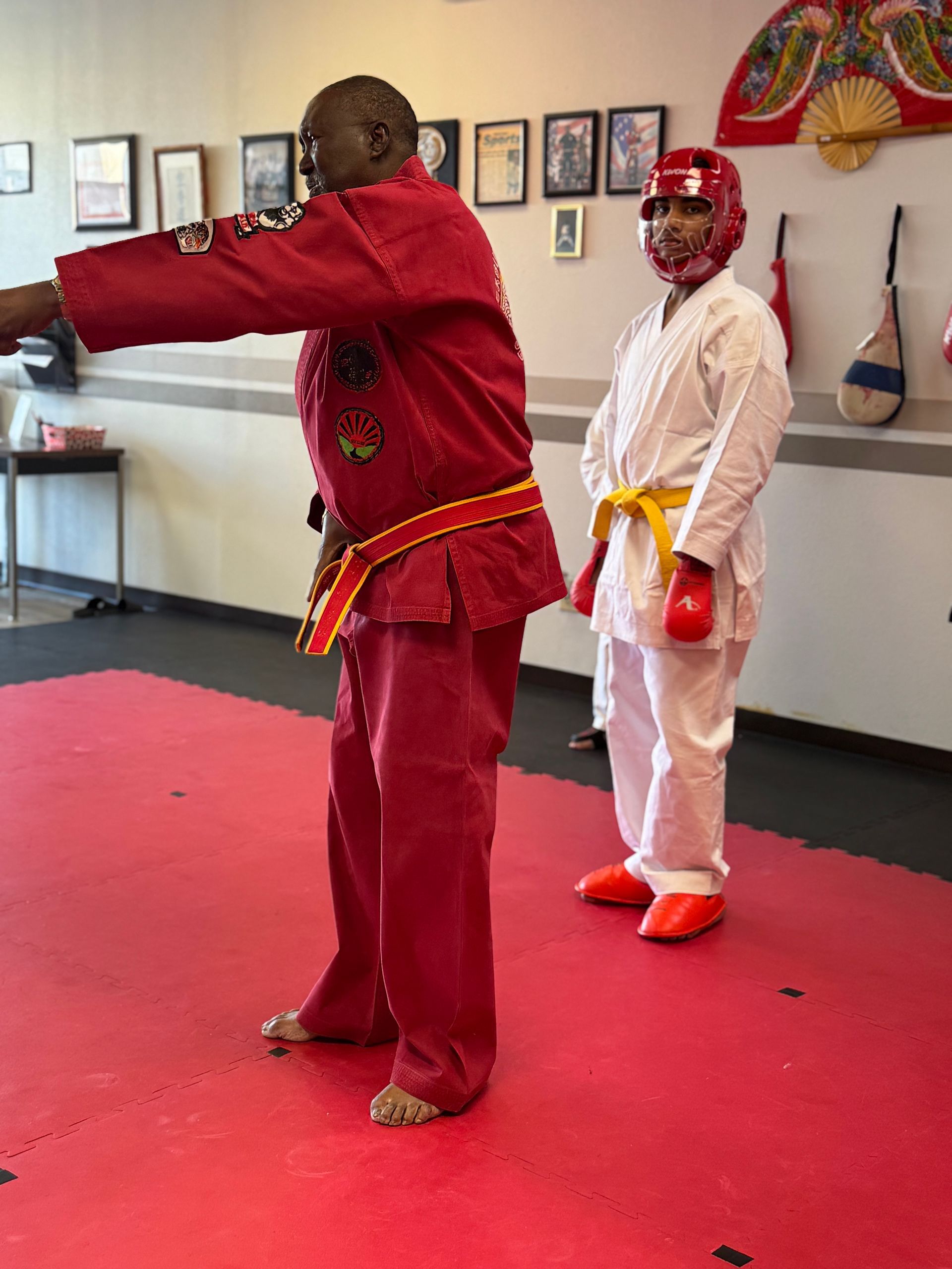 Two martial artists in red and white uniforms practicing a stance indoors on a mat
