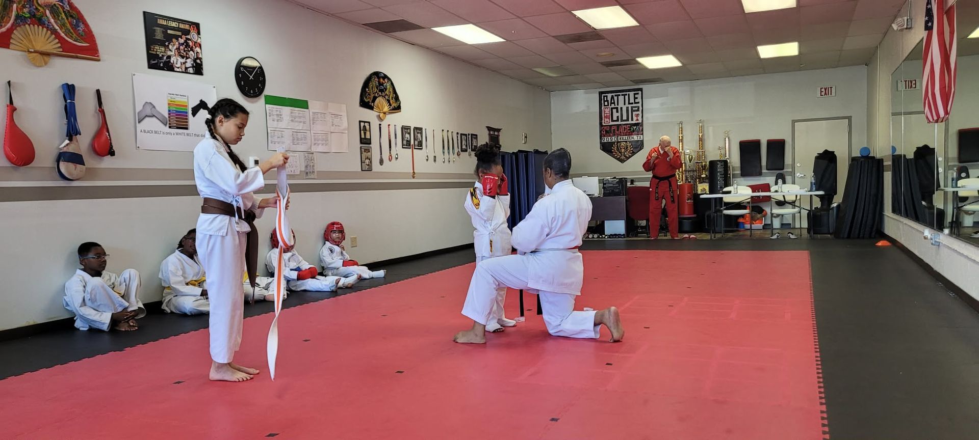 Martial arts class in a studio, with students practicing on a red mat and a teacher instructing.