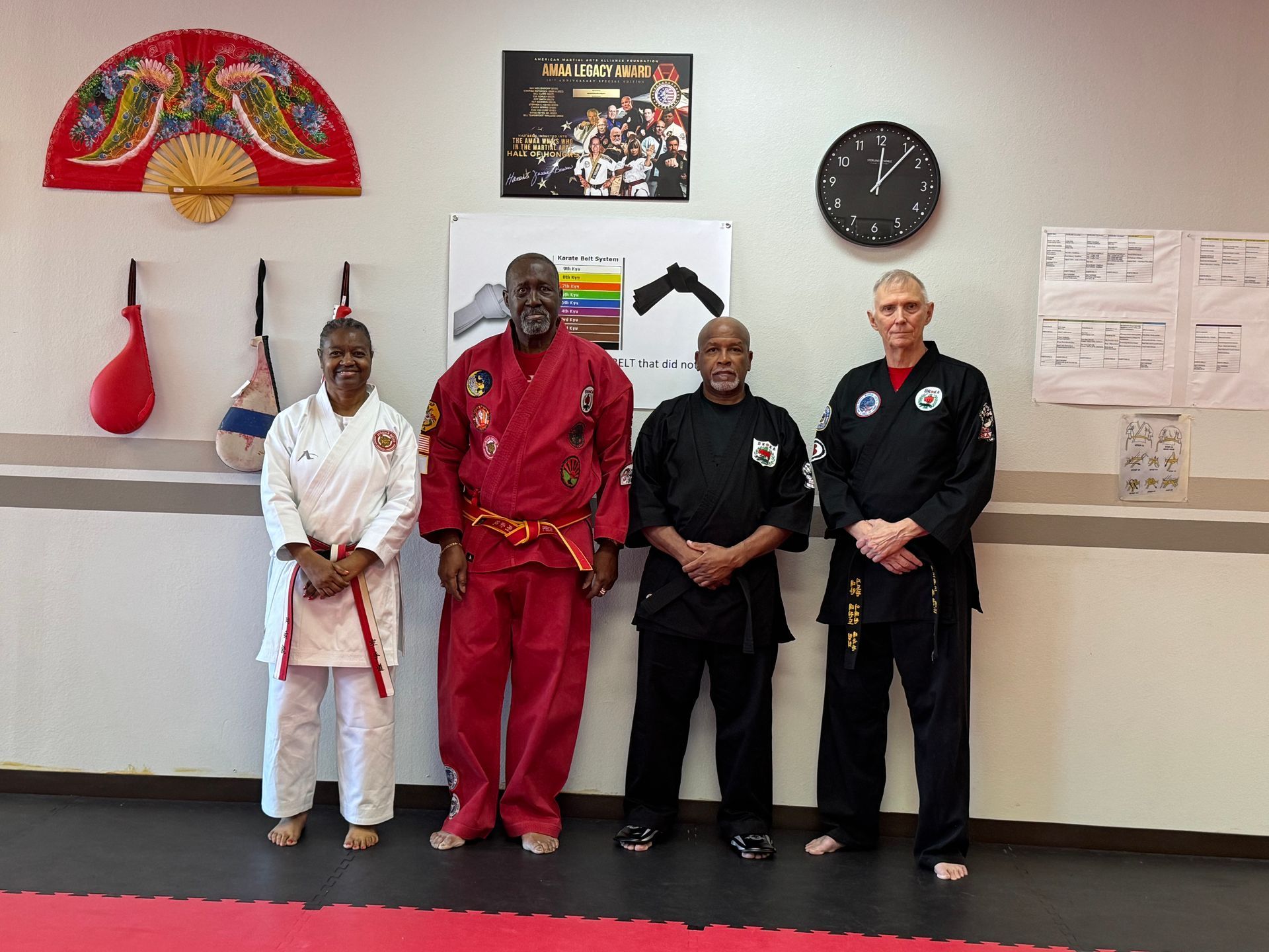 Four martial arts students in uniforms stand in a dojo, holding belts and posing beneath wall displays.