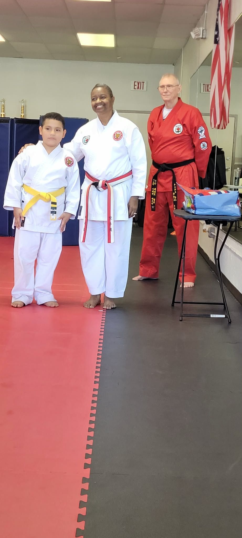 Three martial arts students in gis posing on a dojo mat, with an instructor in red beside them.