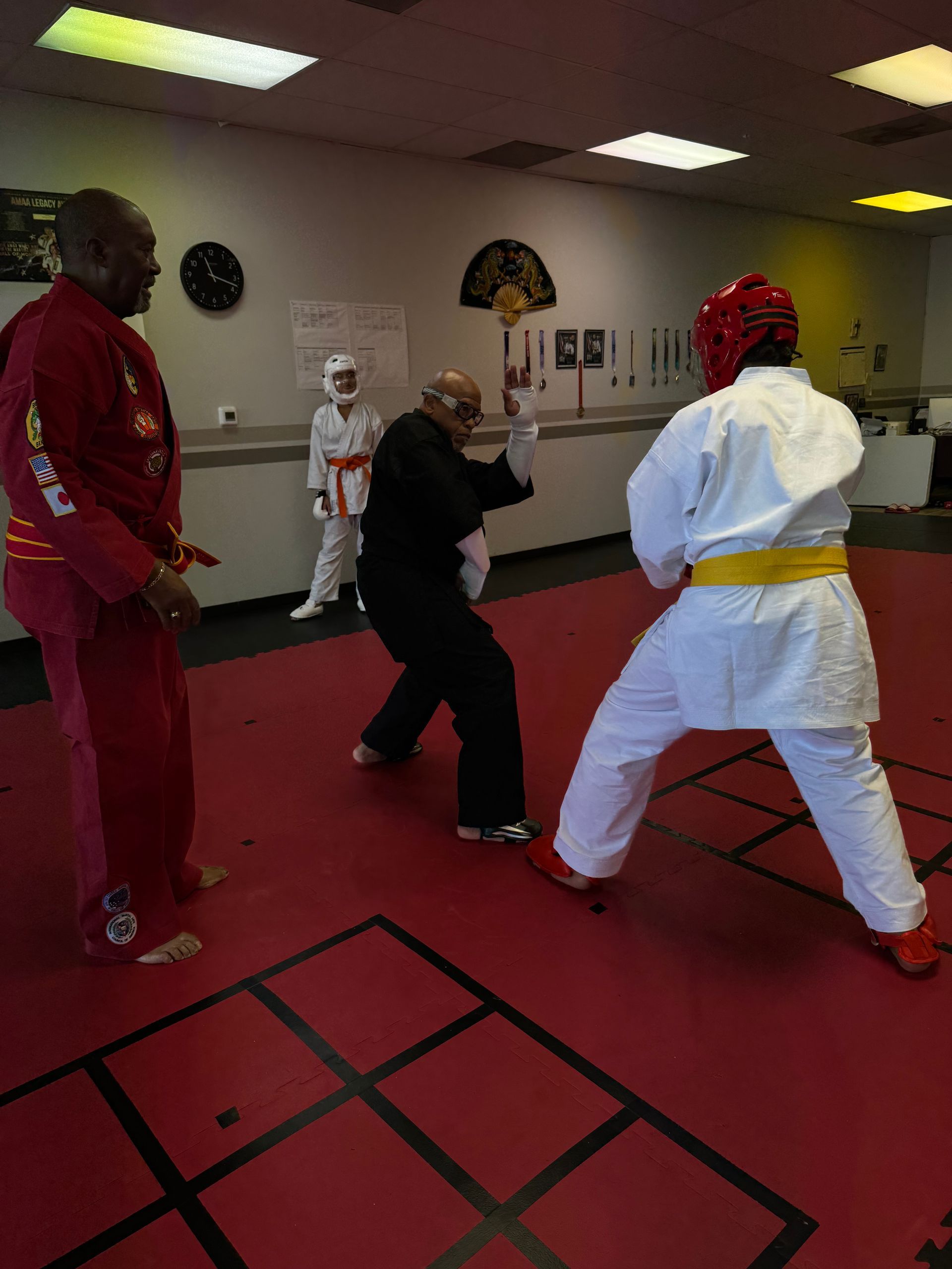 People practicing martial arts in a red-carpeted training room, sparring with protective gear.