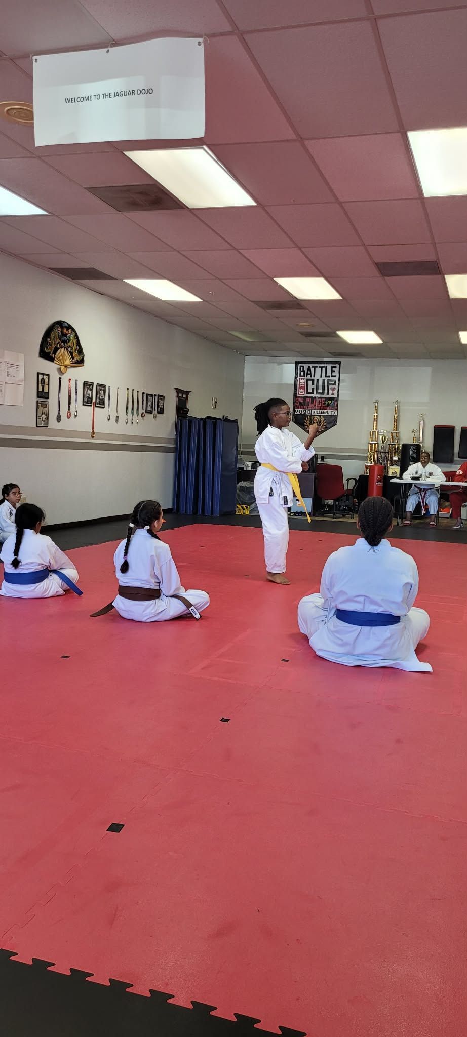 Children in white martial arts uniforms sitting on a red mat in a dojo during class