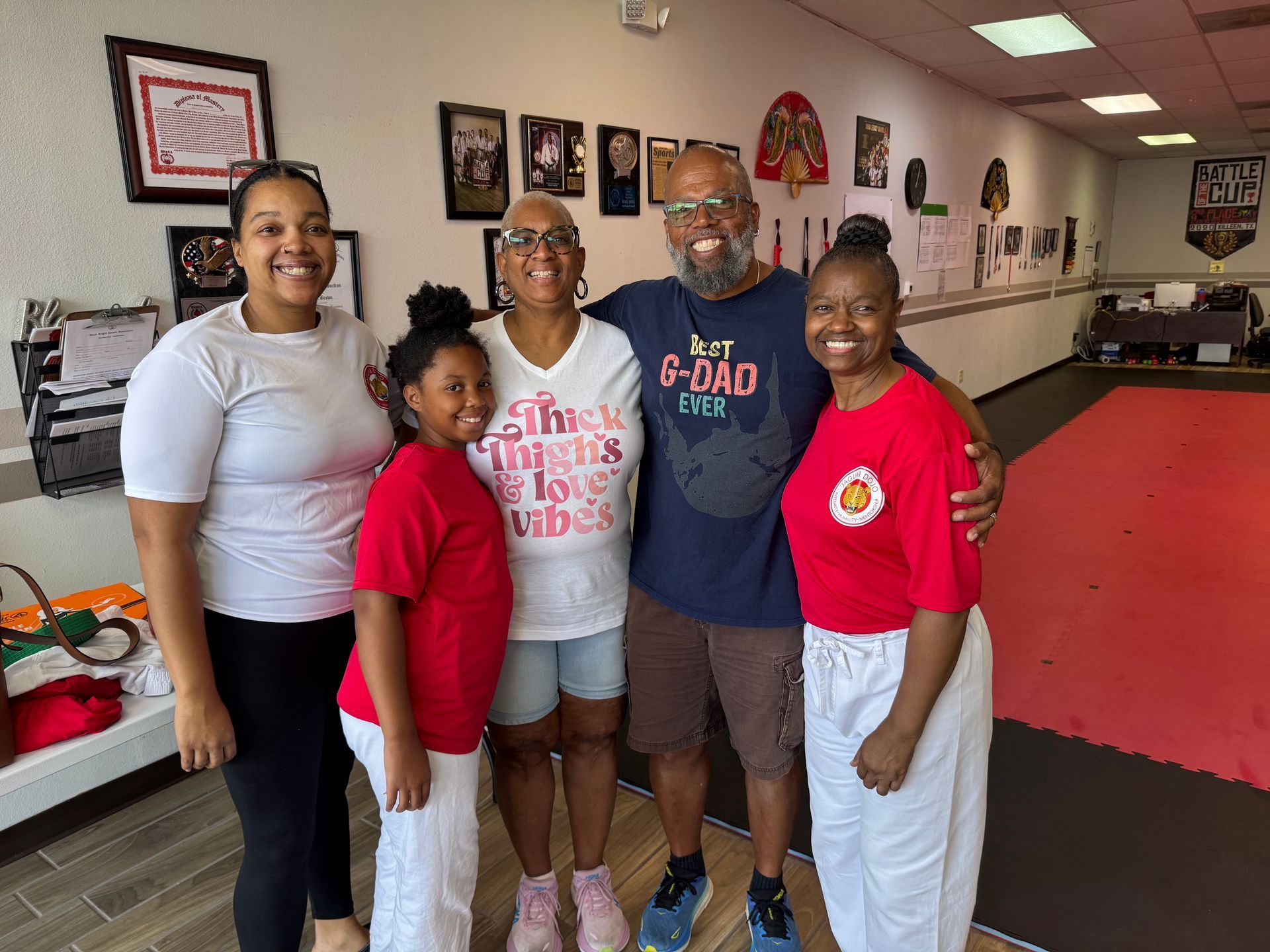 Five people smiling in a community hall with framed photos and a red floor mat.