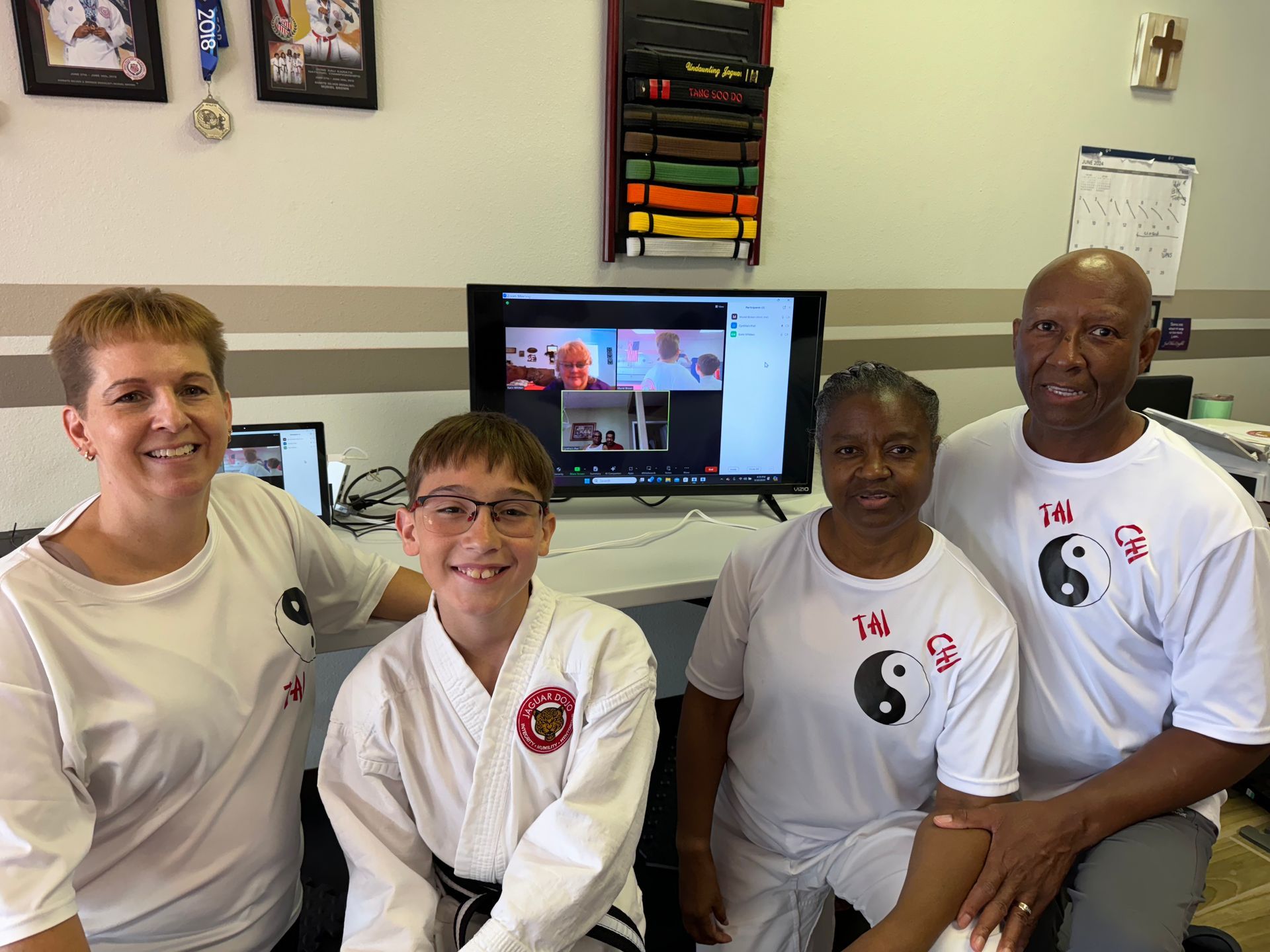 Four people in white shirts smile in a classroom, seated together with a computer screen behind them.