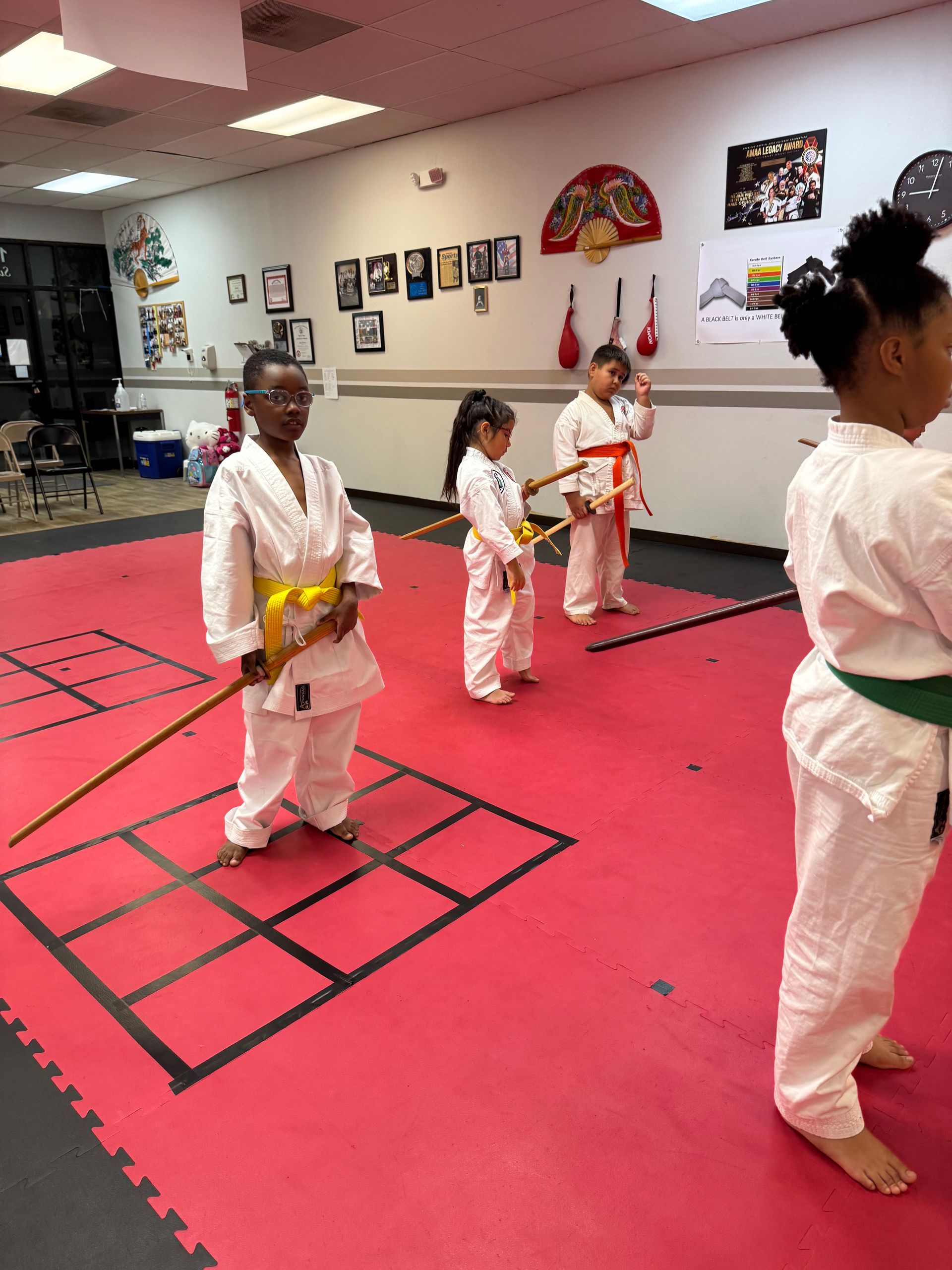 Children in white martial arts uniforms practicing in a red-matted dojo, holding yellow belts