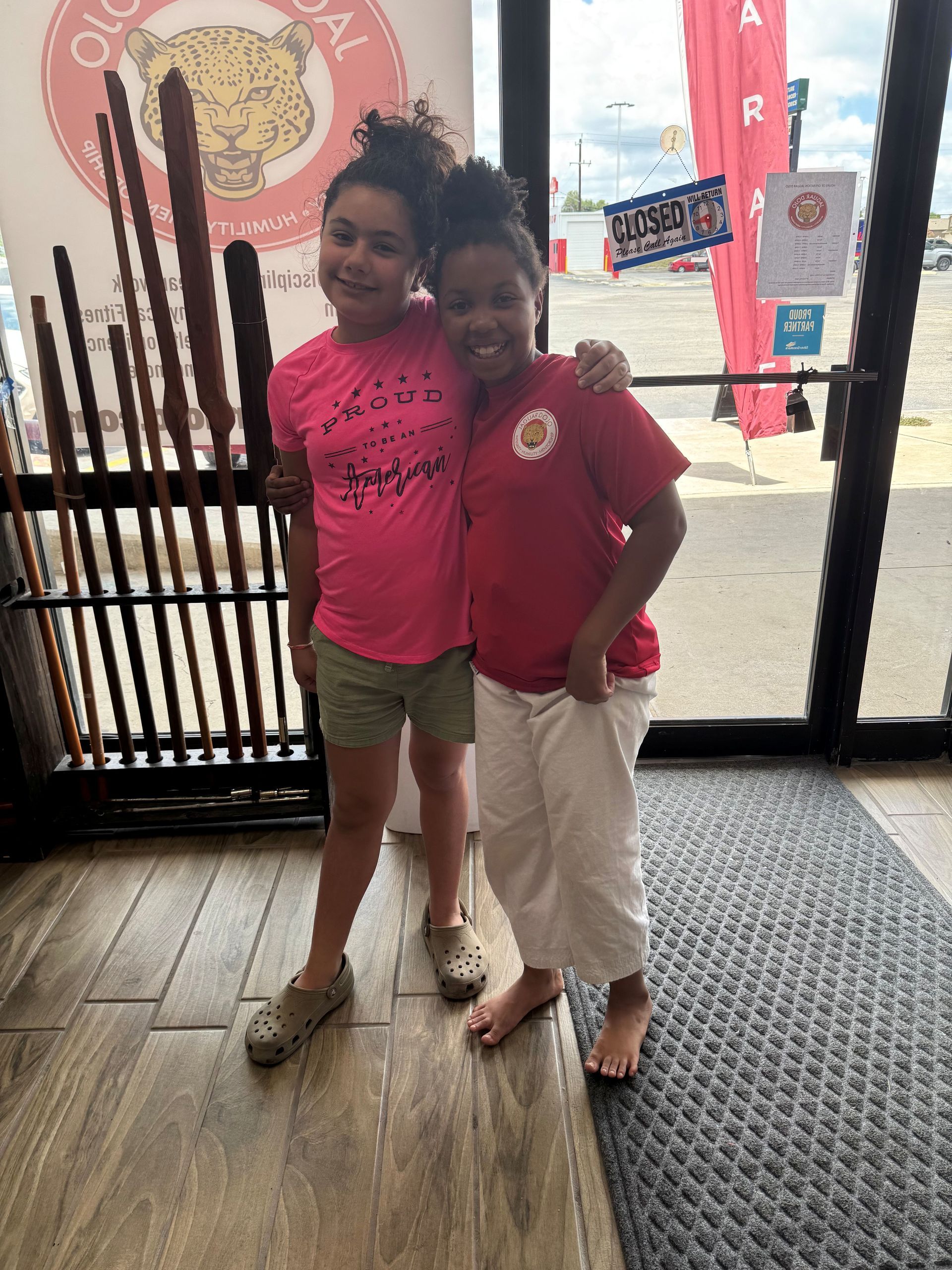 Two children standing together in a storefront doorway, wearing pink and red shirts.
