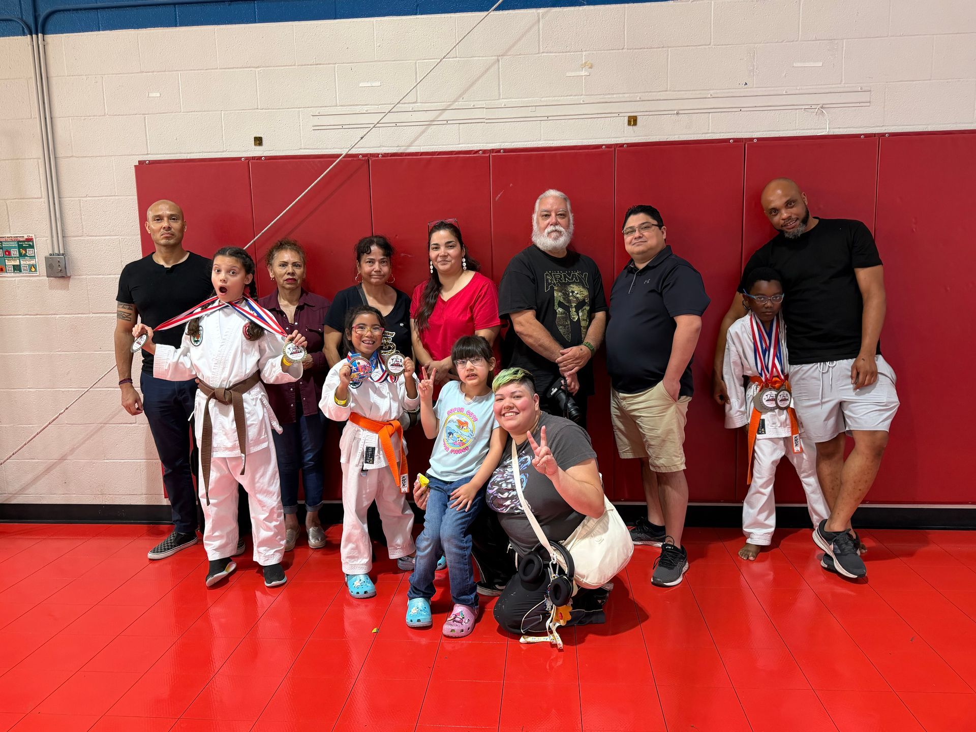 Group posing in a gym, some in martial arts uniforms, against a red and white wall.