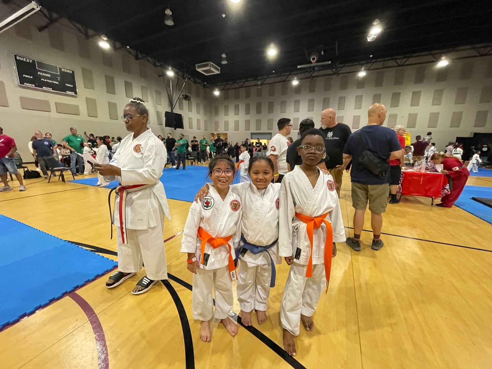Four children in white karate uniforms with orange belts stand in a gymnasium during a martial arts event