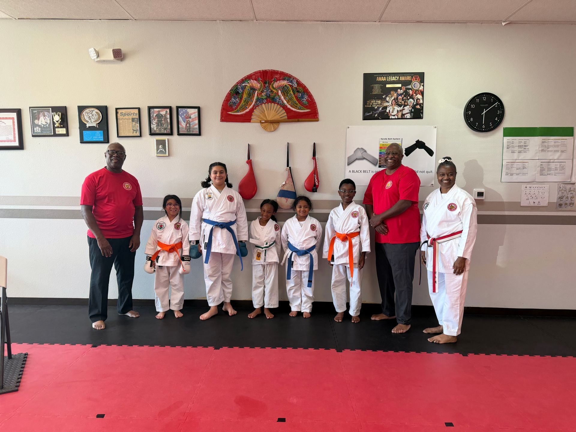 Martial arts class posing in a dojo with black, red, and white uniforms on a red mat.