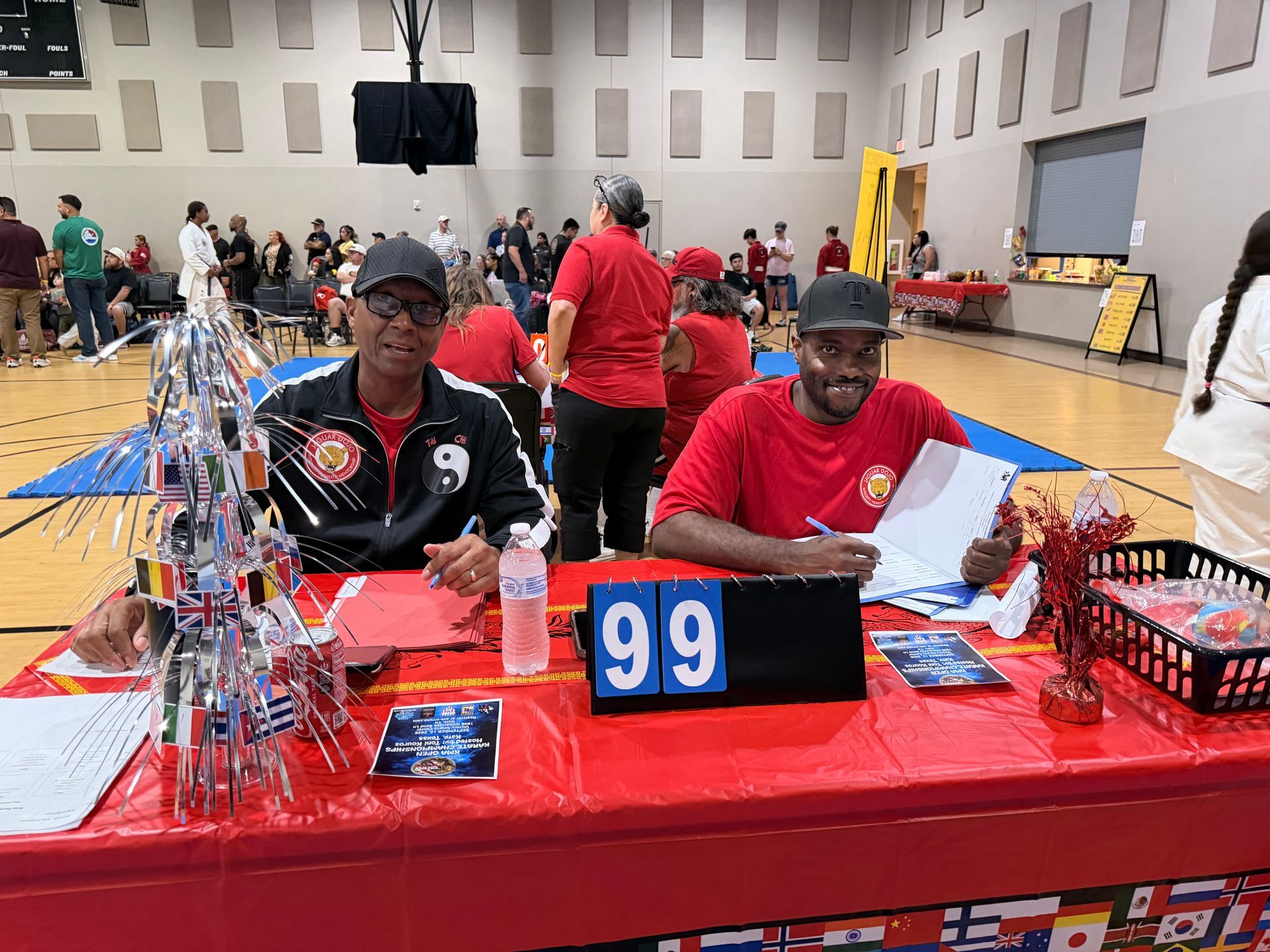 Two people at a red table labeled 99 at an indoor event, with trophies and display items.