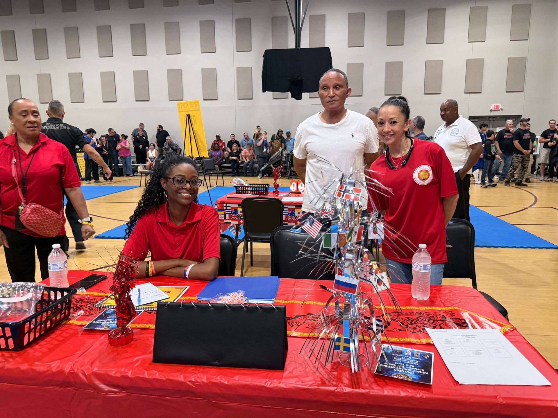 People at a red table in a gymnasium, with a trophy display and event banners in the background.