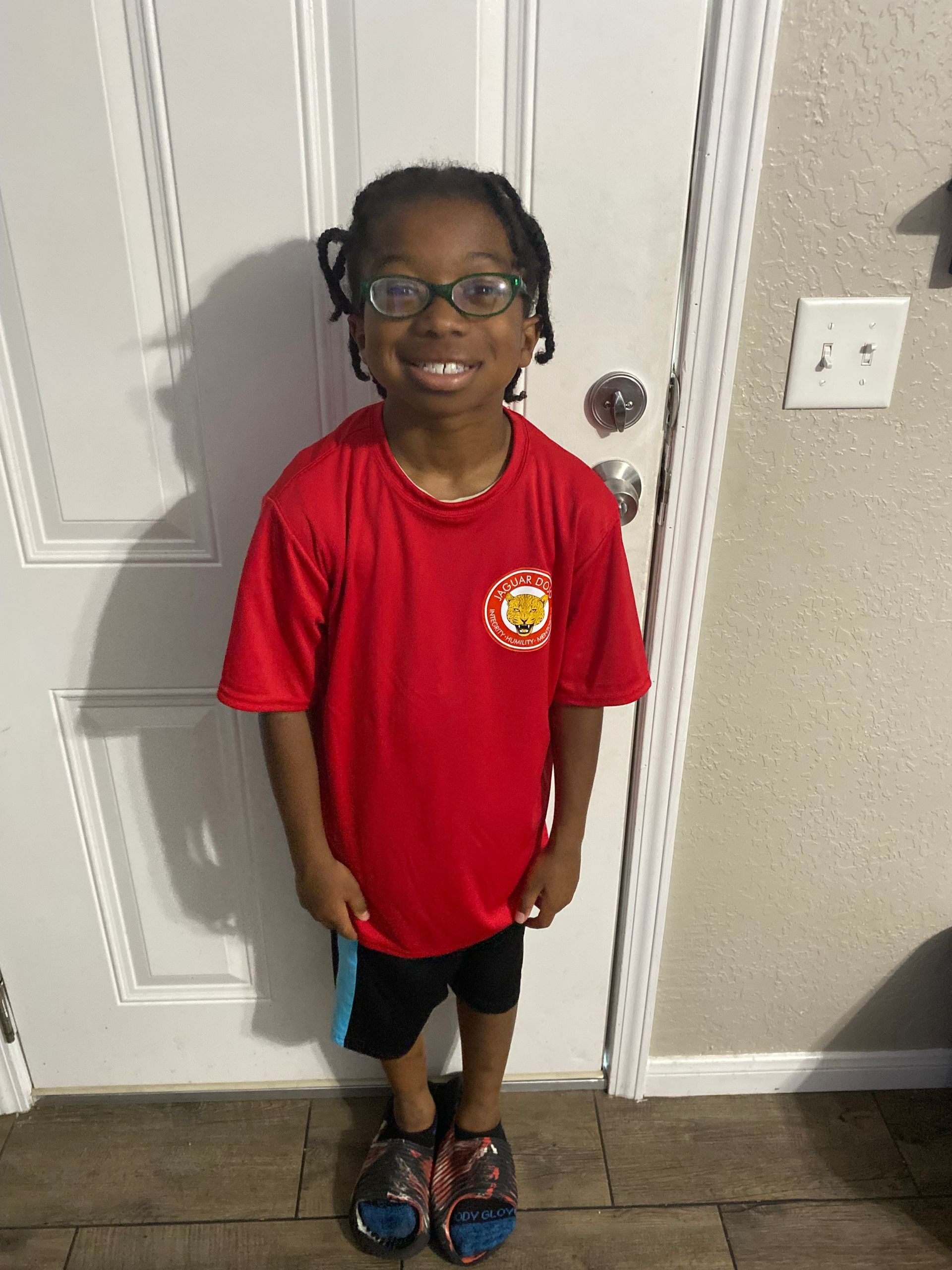 Child in red shirt and black shorts standing indoors by a white door, smiling at the camera.