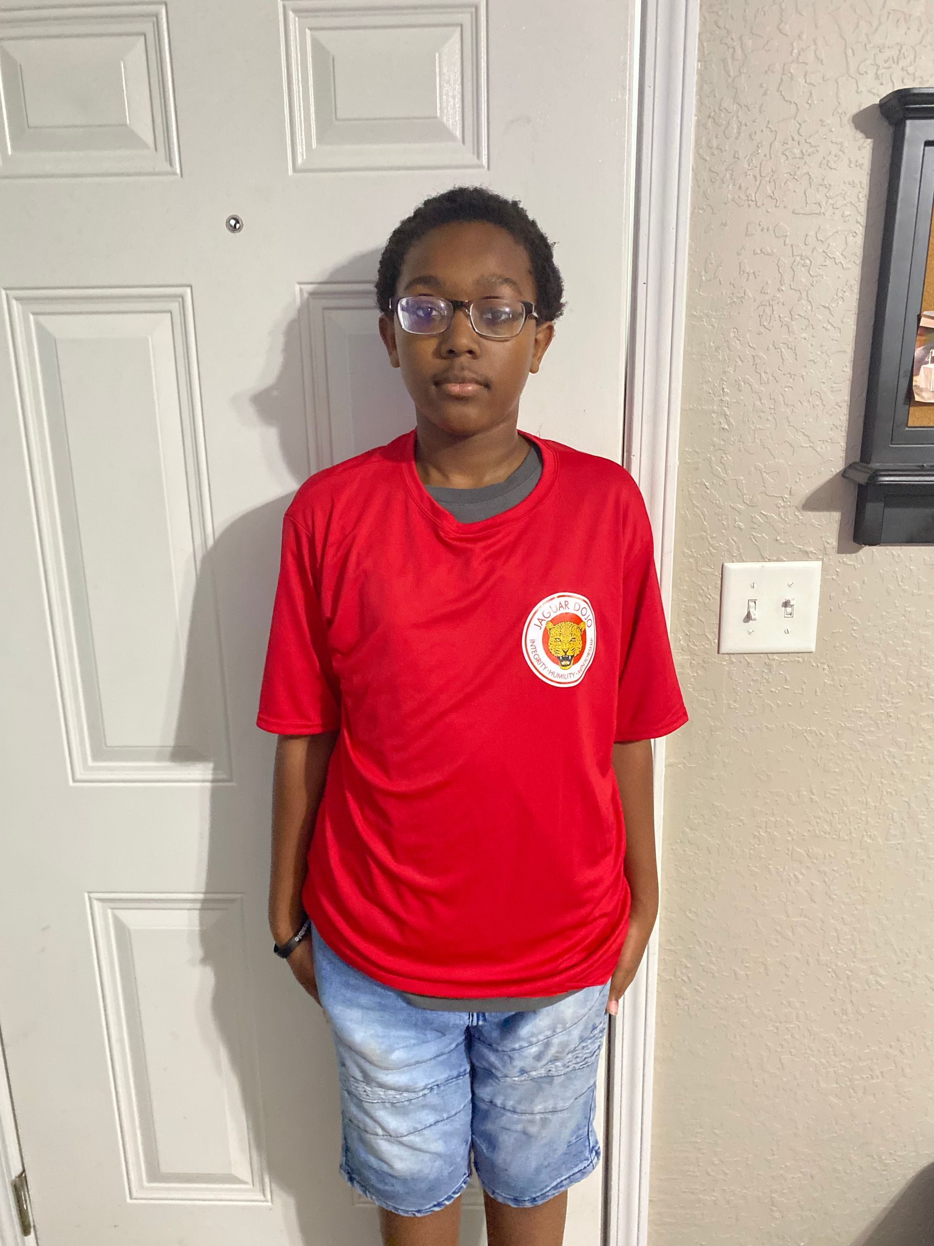Person in a red T-shirt and denim shorts standing indoors by a white door.