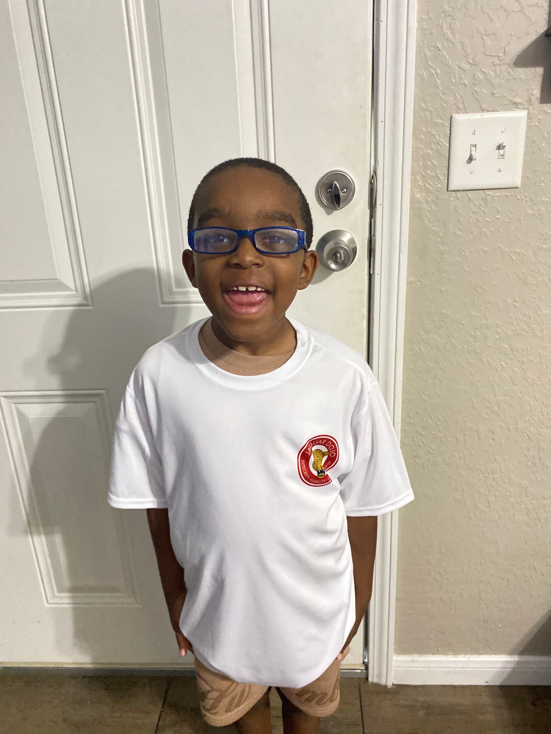 Smiling child in a white T-shirt and blue glasses standing by a white door.