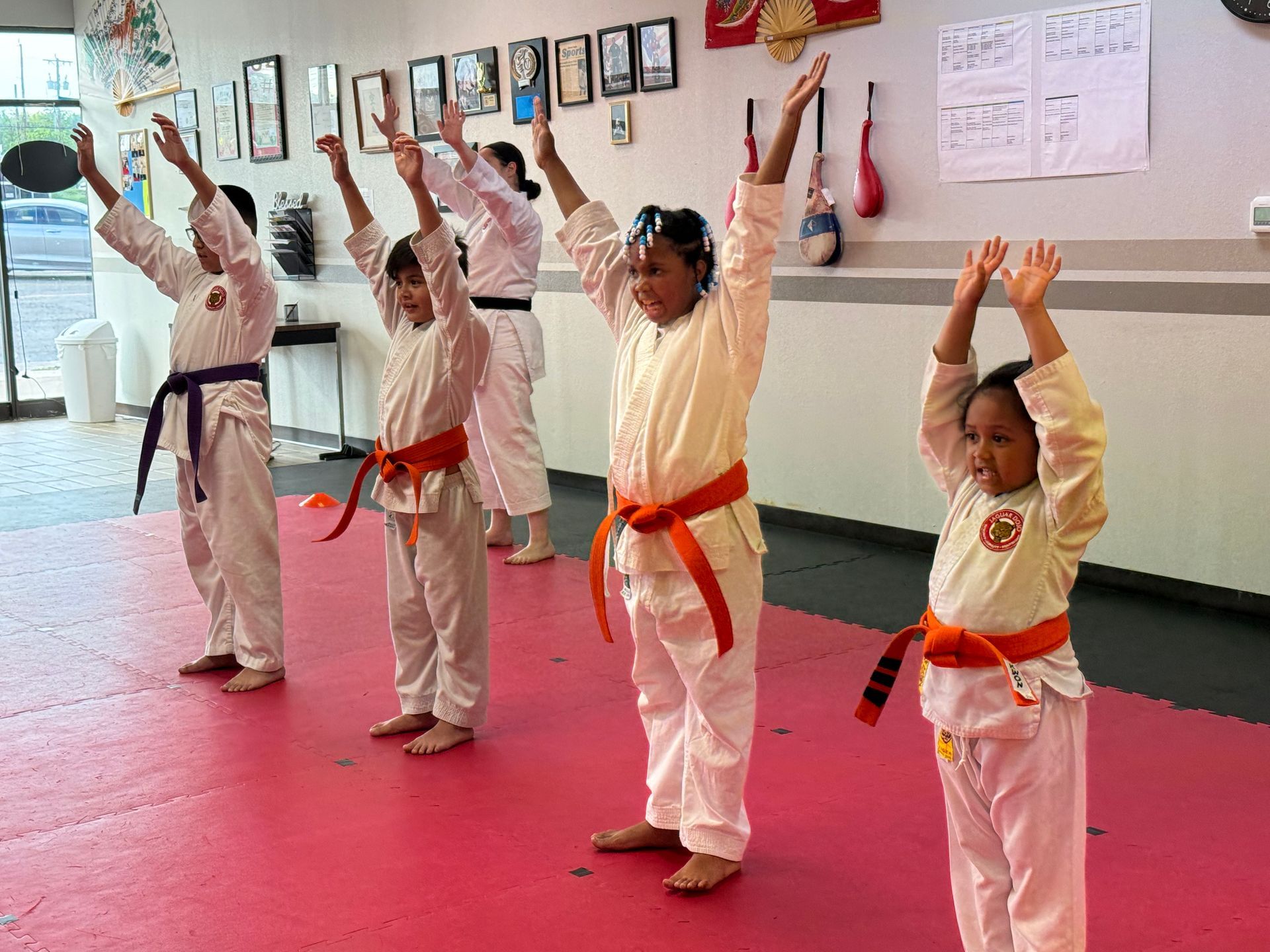 Children in white martial arts uniforms practicing a raised-arm pose in a dojo