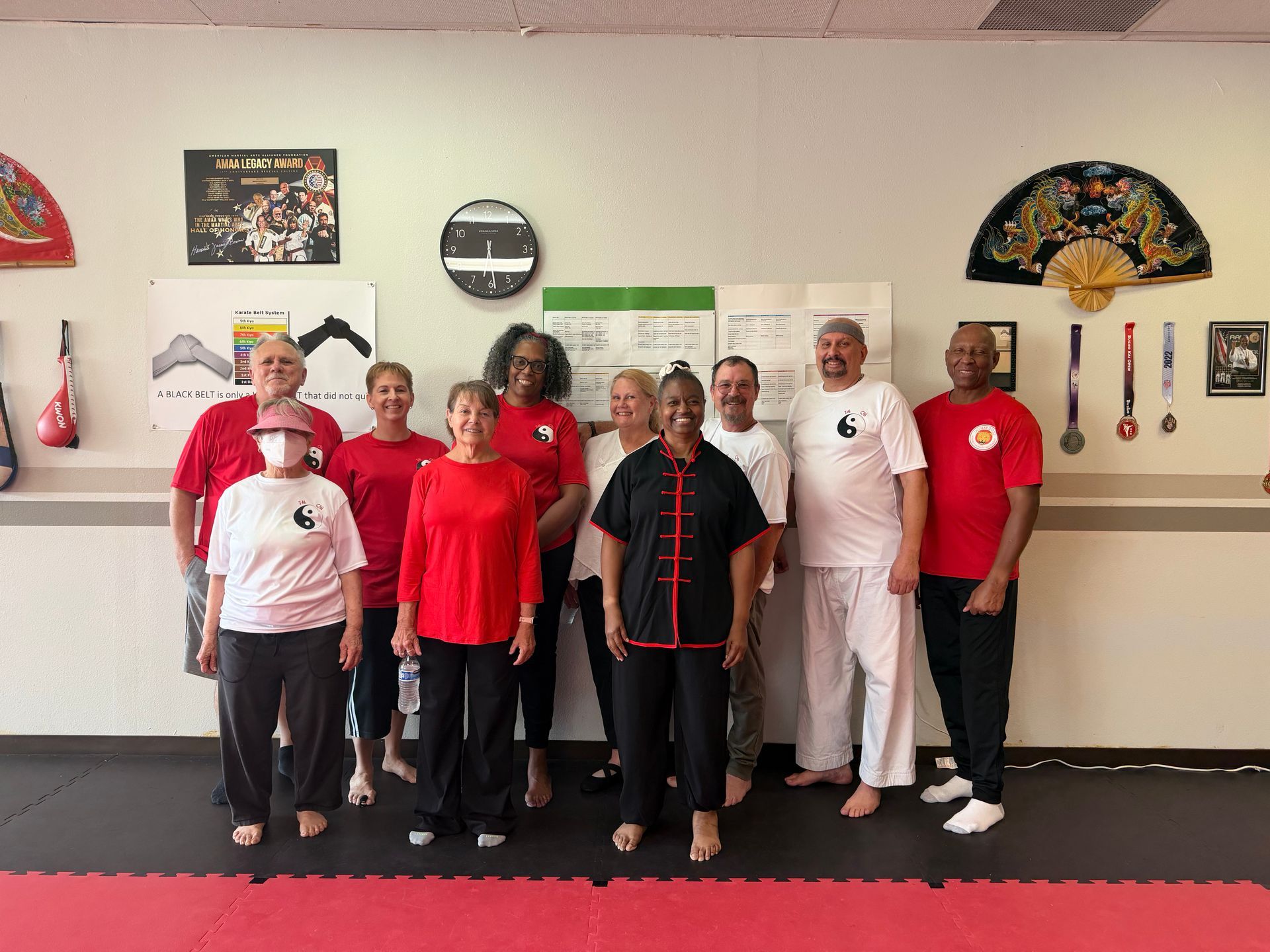 Group of people in red and white shirts posing in a martial arts studio with fans and framed decor on the wall