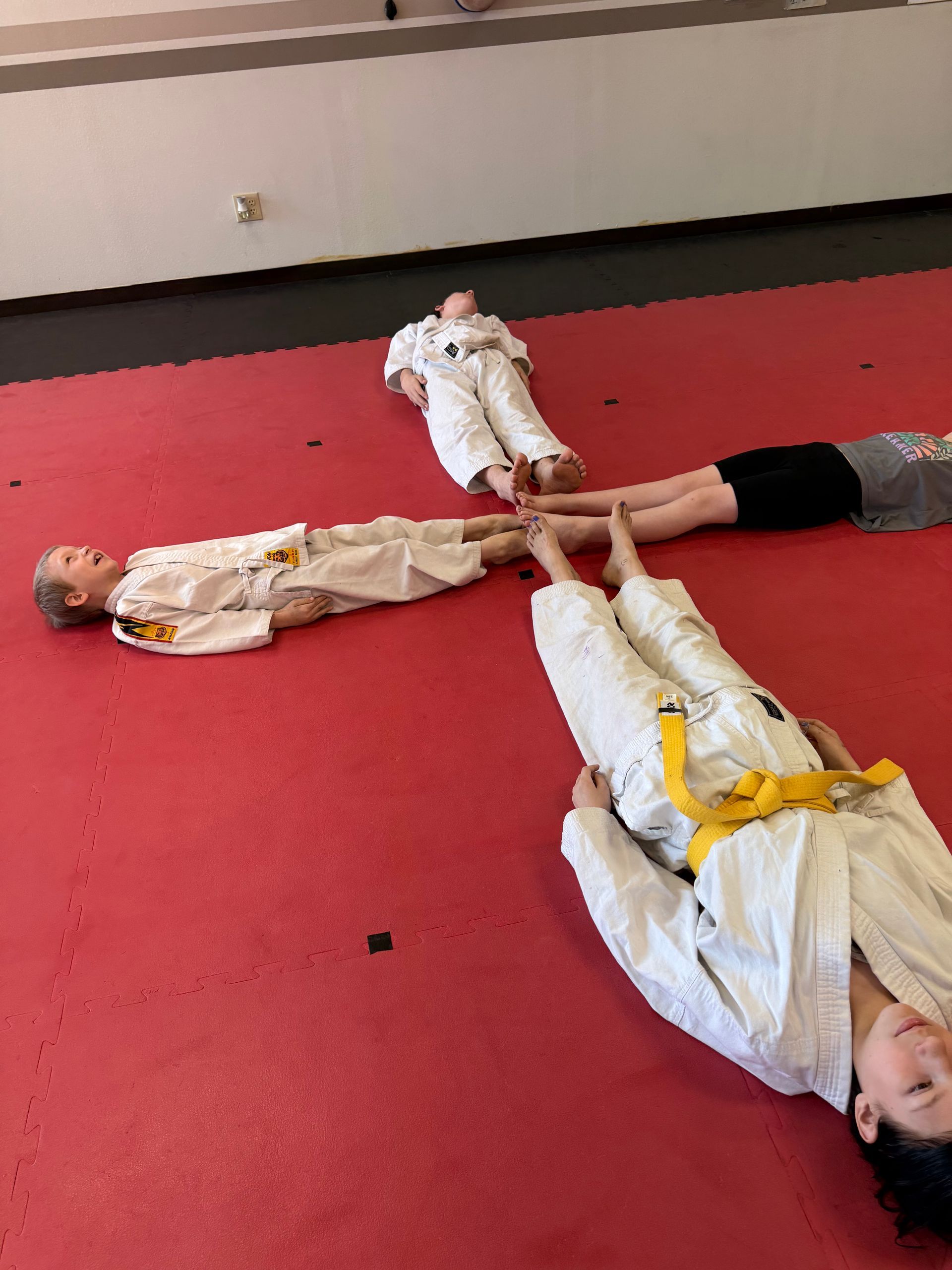 Children in white costumes lie on a red mat, feet joined together in a circle.