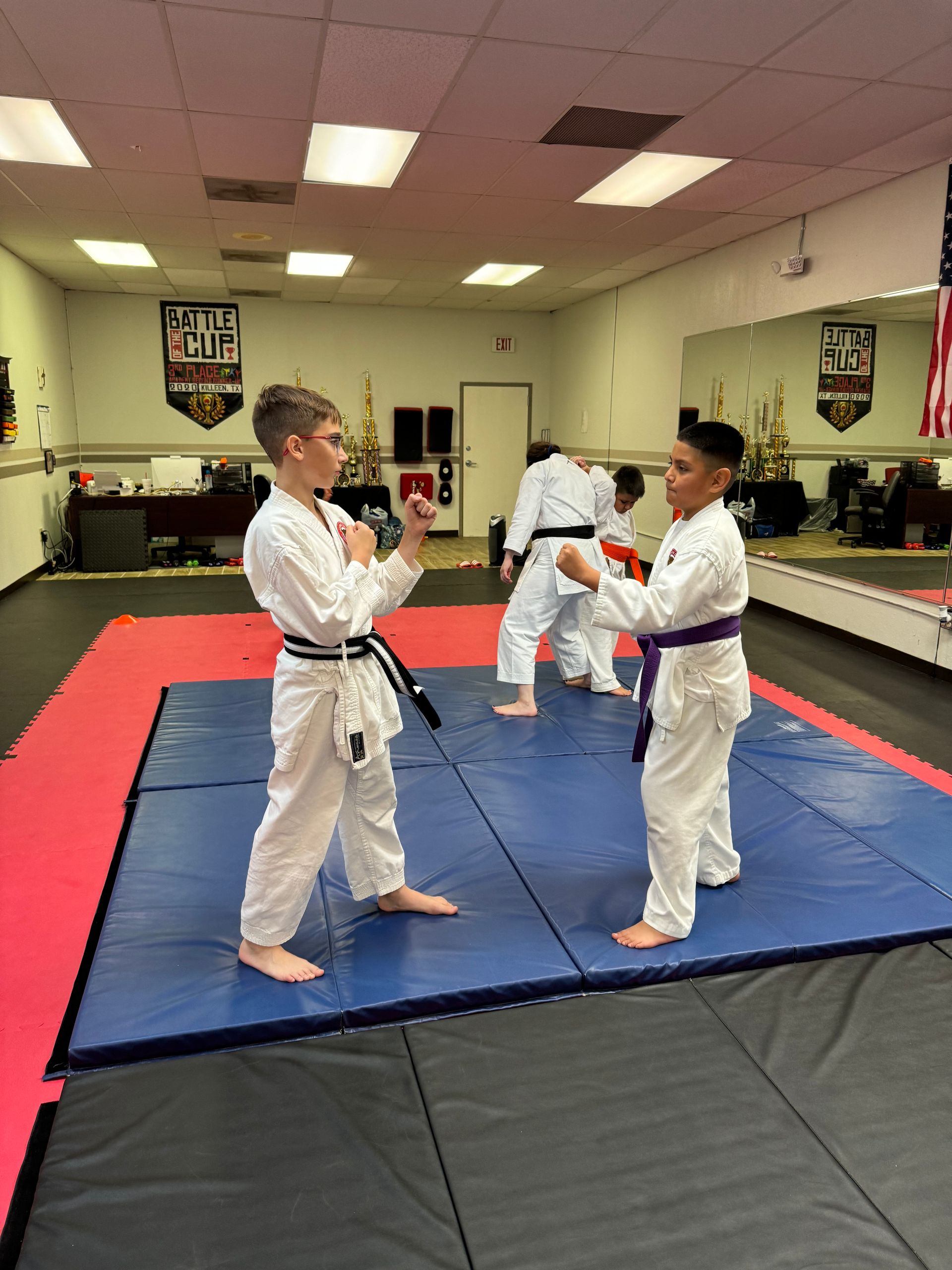 Three martial artists practice sparring on red and blue mats in a dojo.