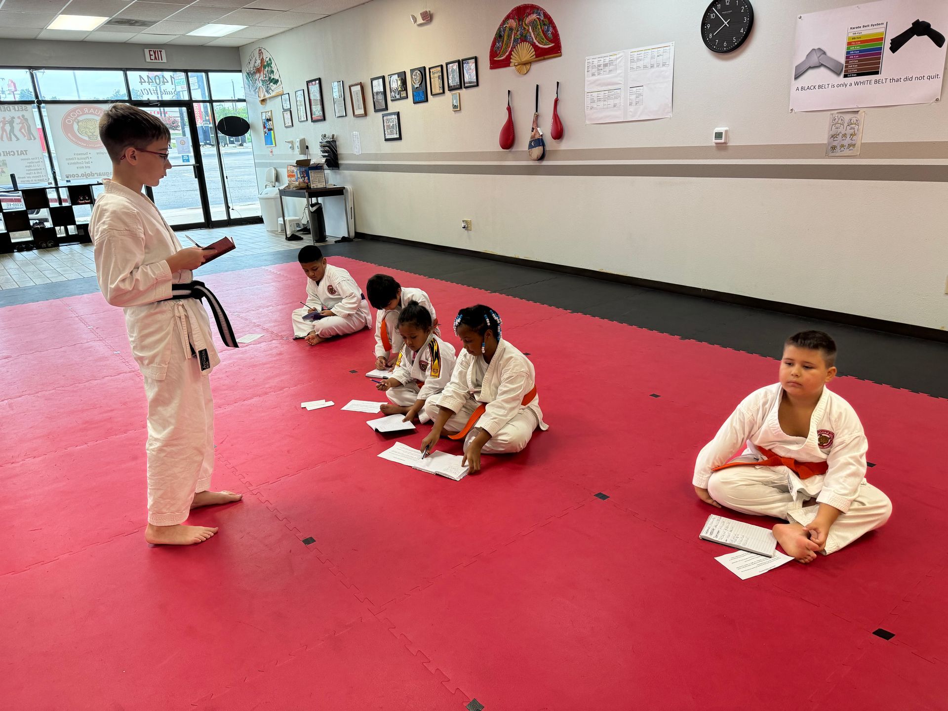 Children in white martial arts uniforms sitting on red mats in a dojo while an instructor stands nearby