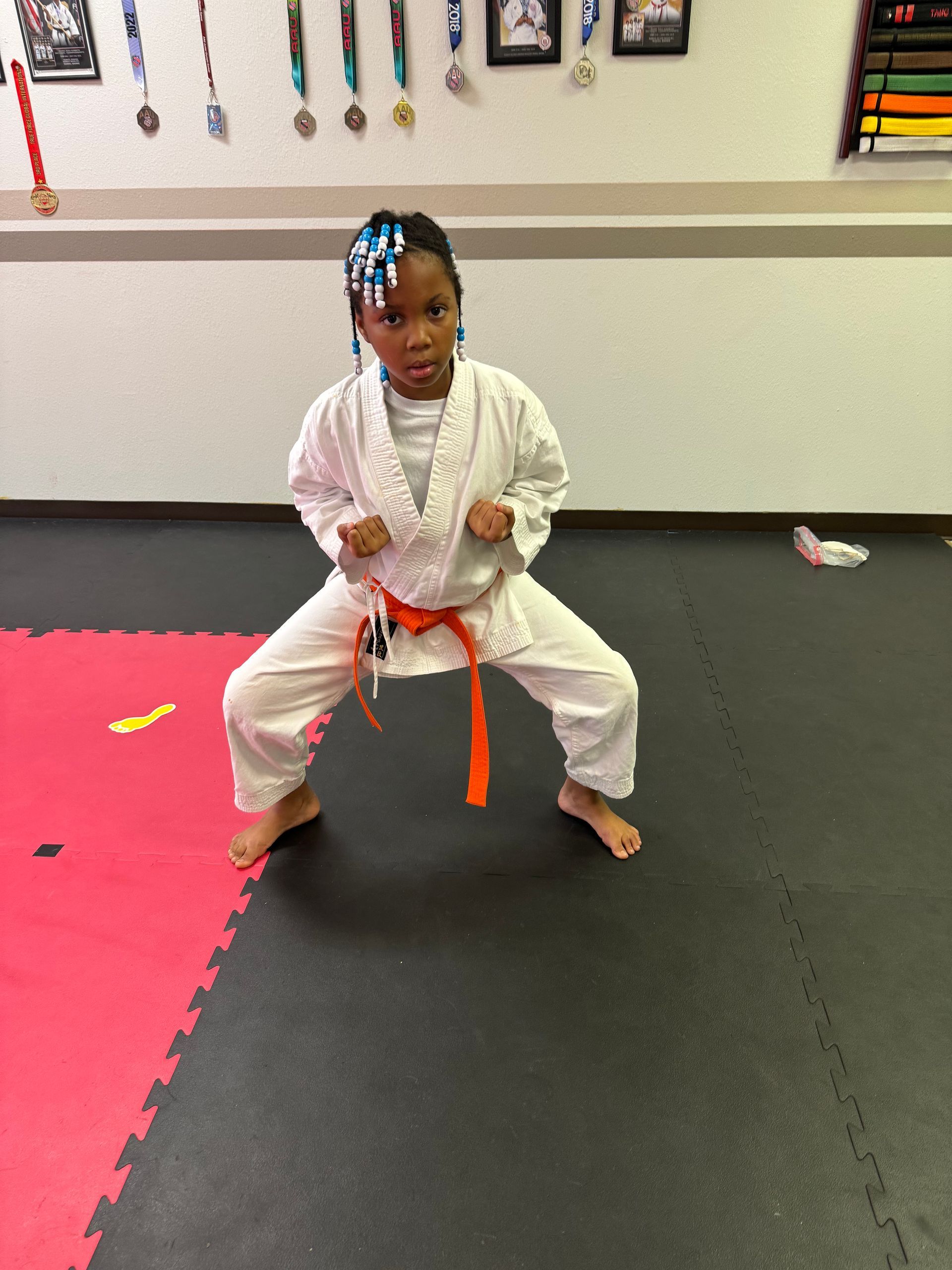 Child in a white martial arts uniform and orange belt in a low stance on padded mats.