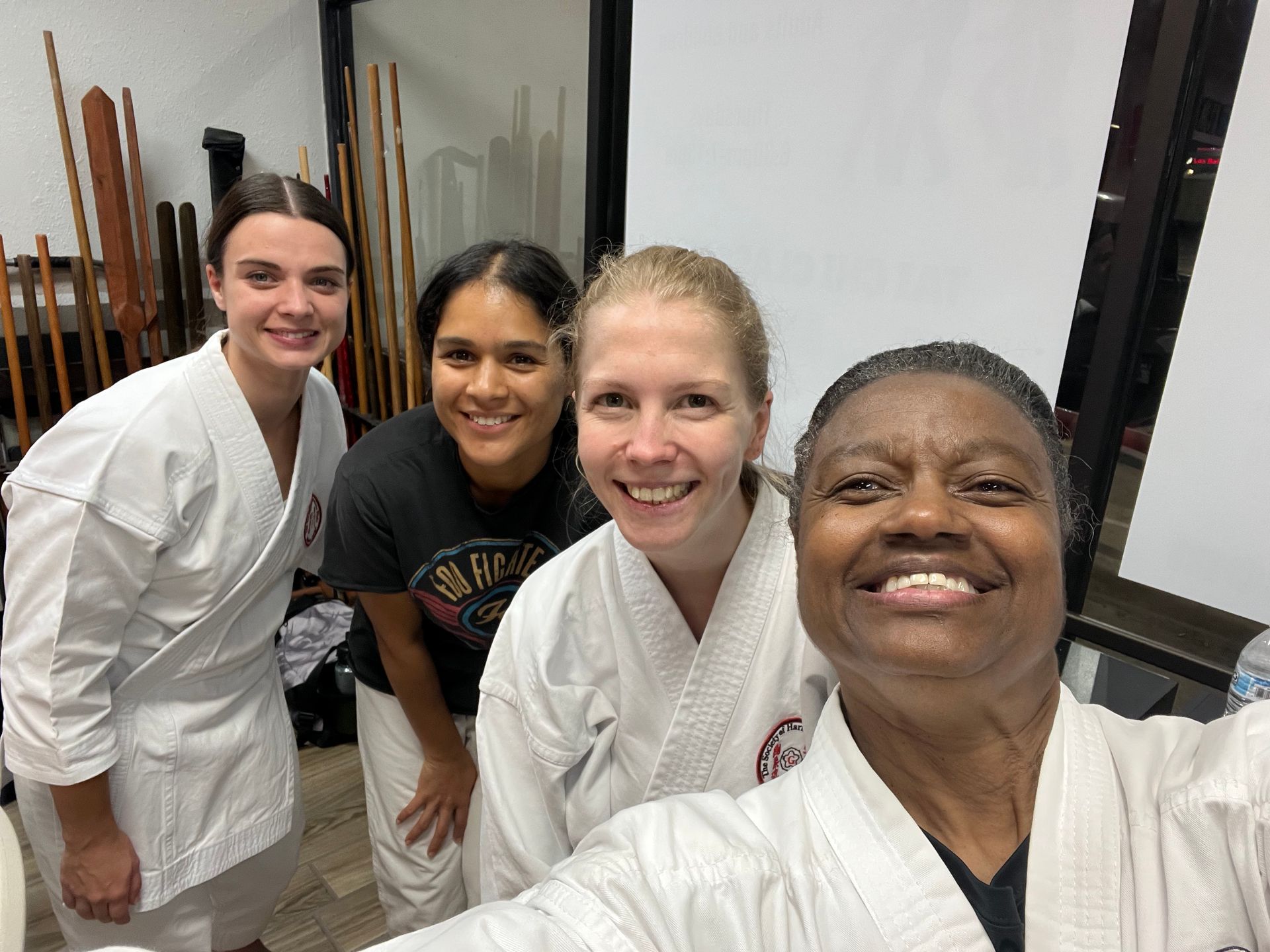 Four people in white martial arts uniforms smile for a group selfie in a training room.