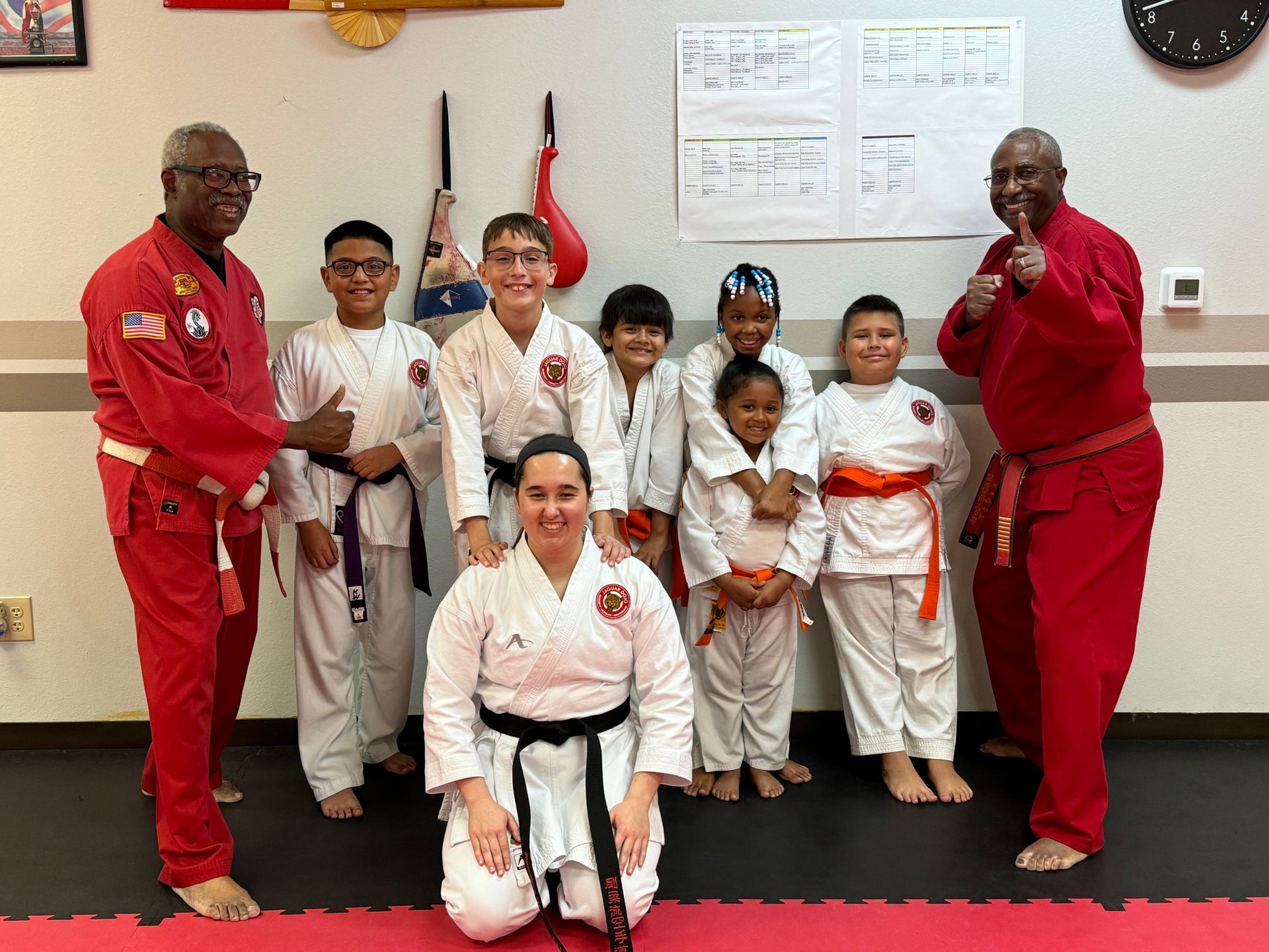 Martial arts class posing in white and red uniforms in a dojo with flags and a wall clock