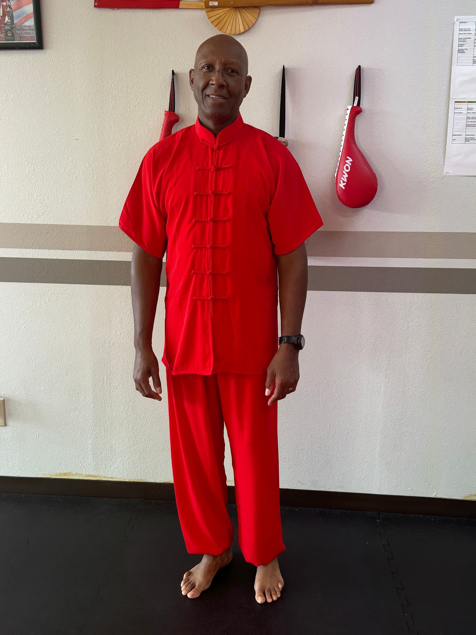 Person in a red martial arts uniform standing barefoot indoors against a wall.