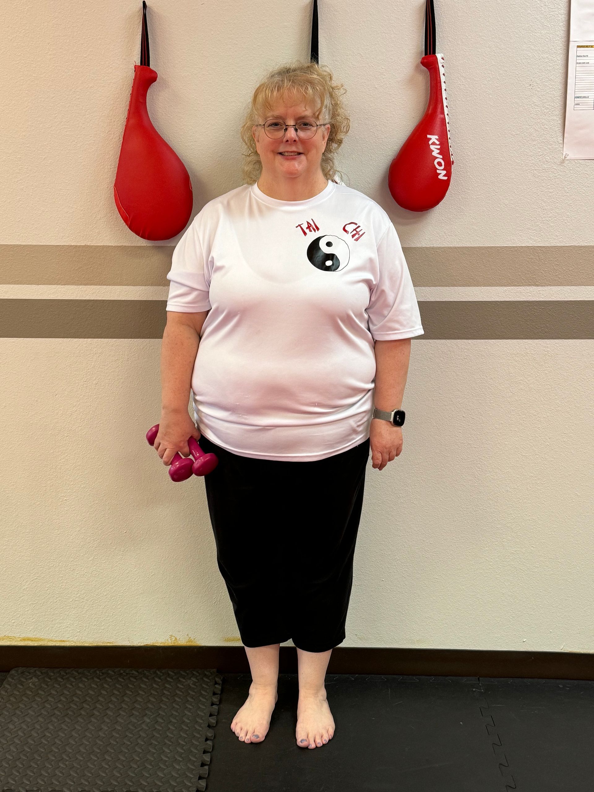 Woman standing barefoot against a wall, holding pink dumbbells, wearing a white T-shirt and black pants.