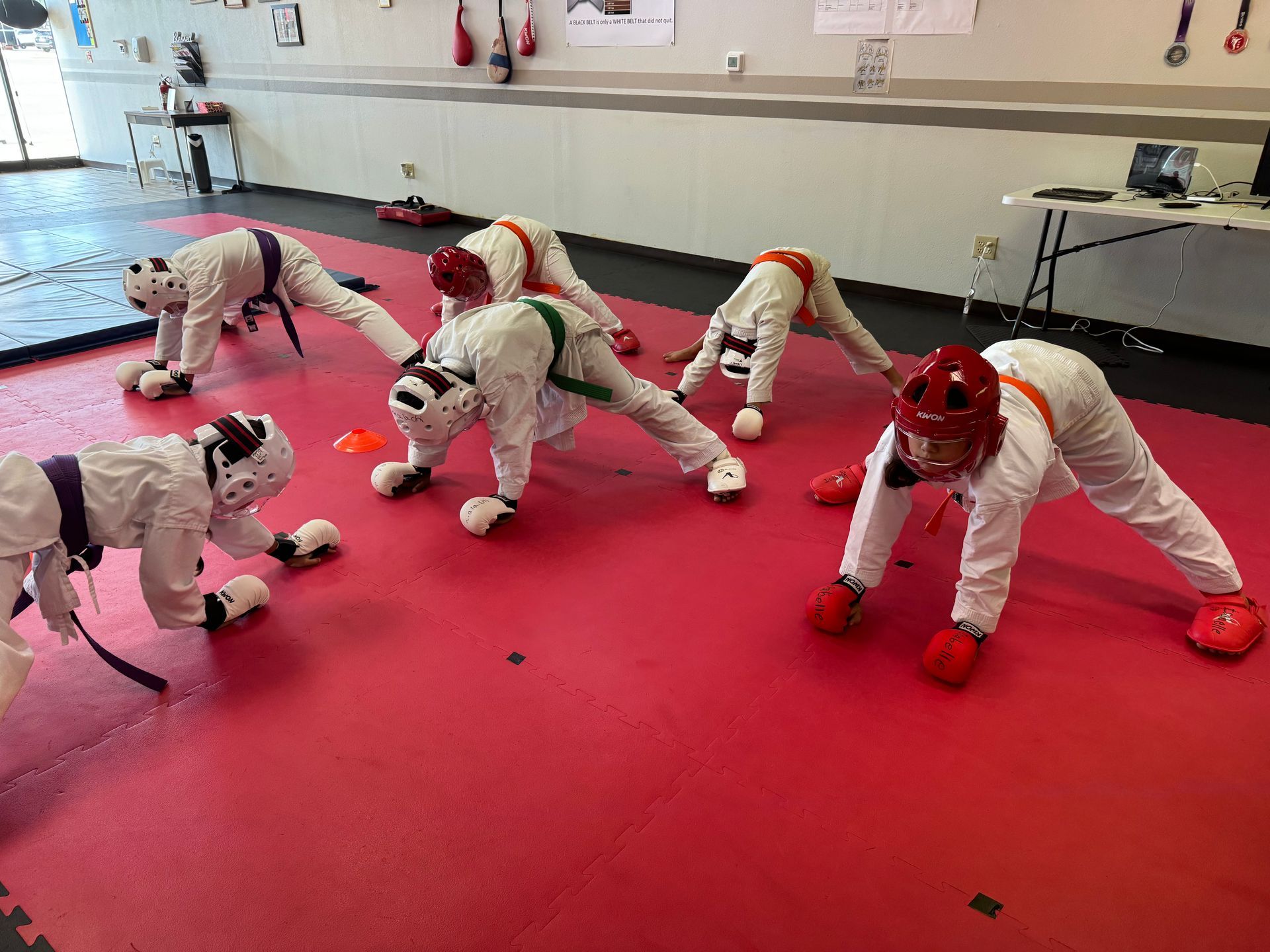 Children in white fencing gear stretching on a red mat in a practice room