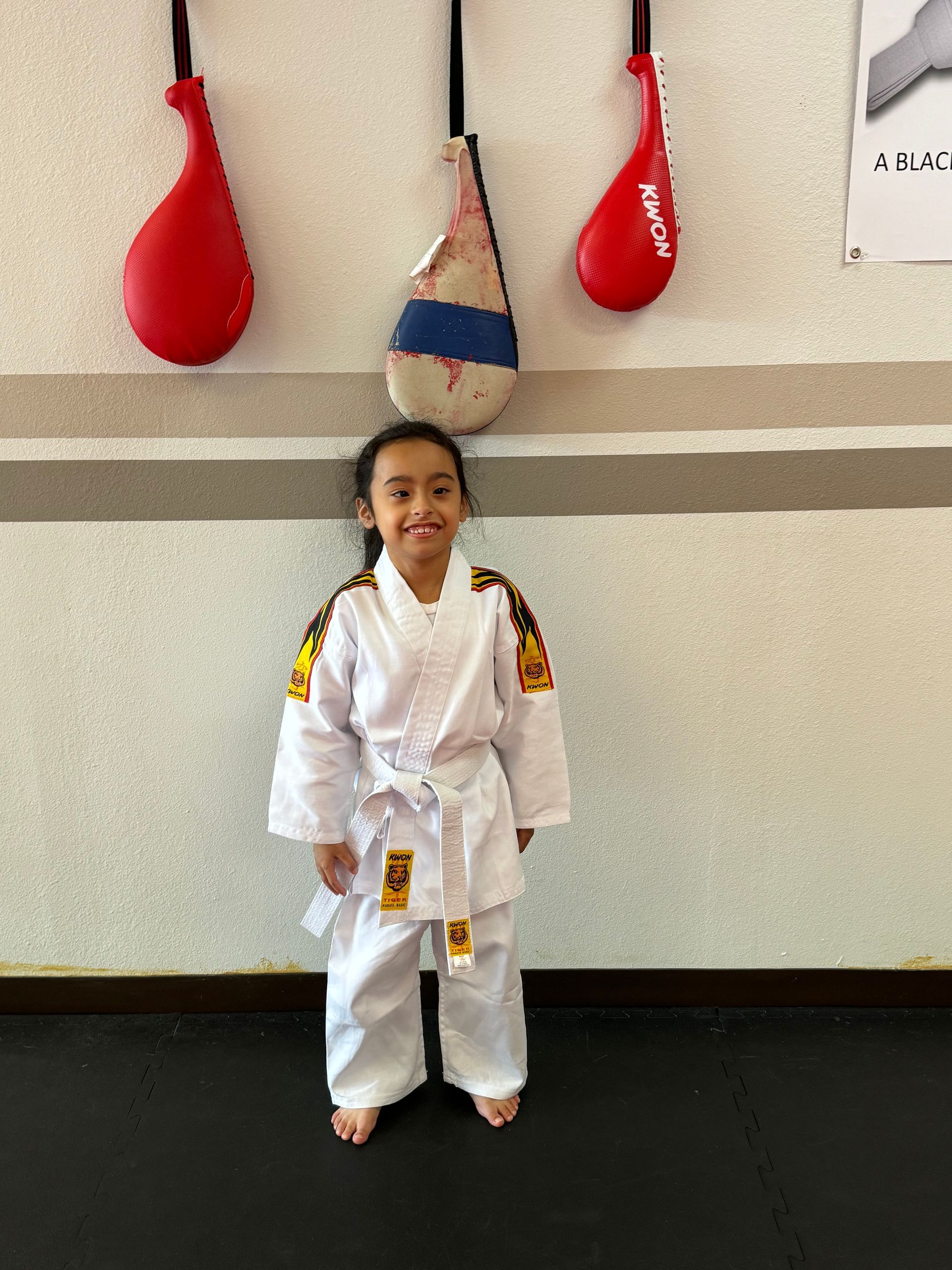 Child in a white martial arts uniform standing indoors in front of hanging red and blue punching bags