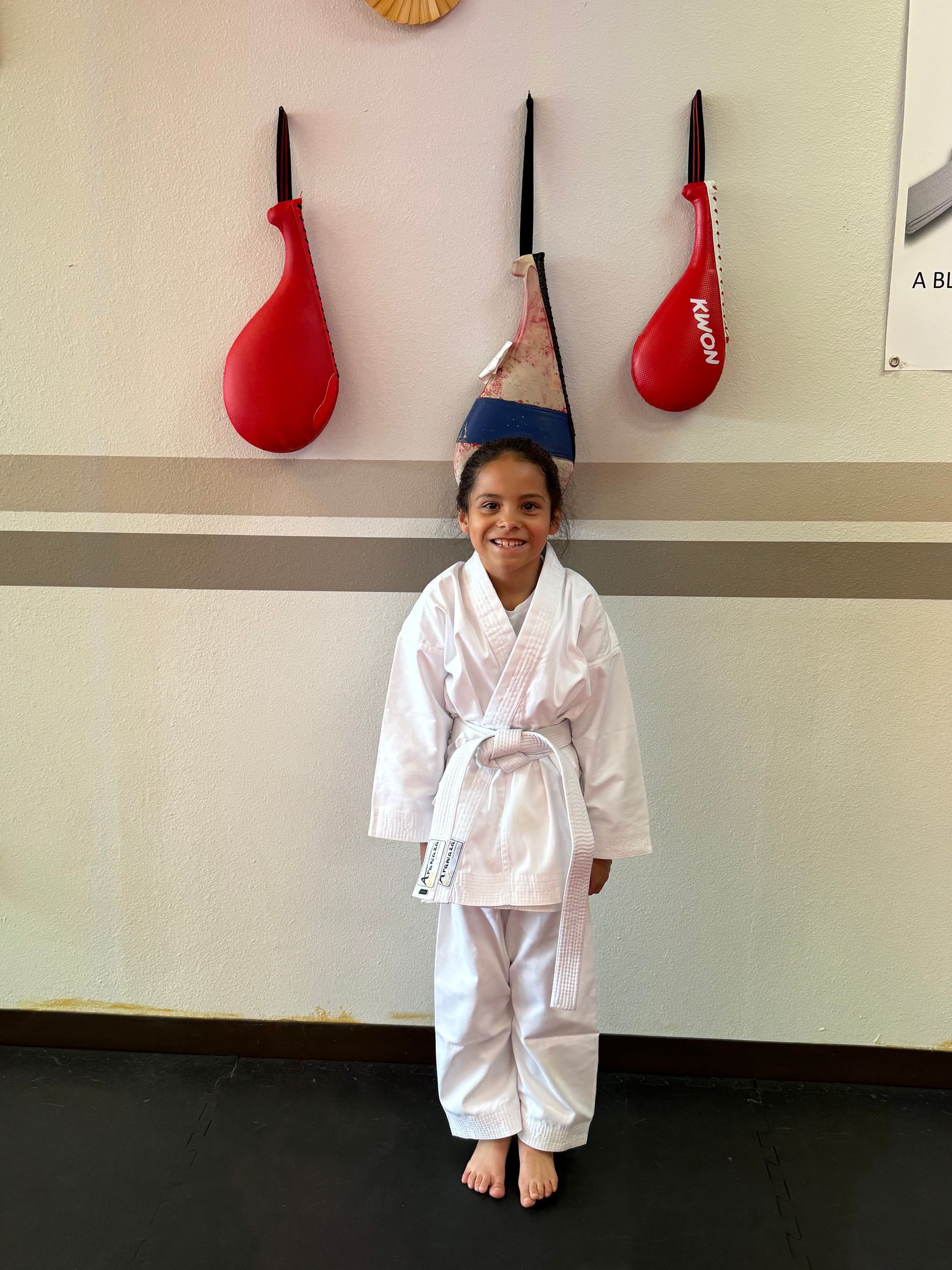 Child in a white martial arts uniform standing indoors beneath hanging red and blue training bags.