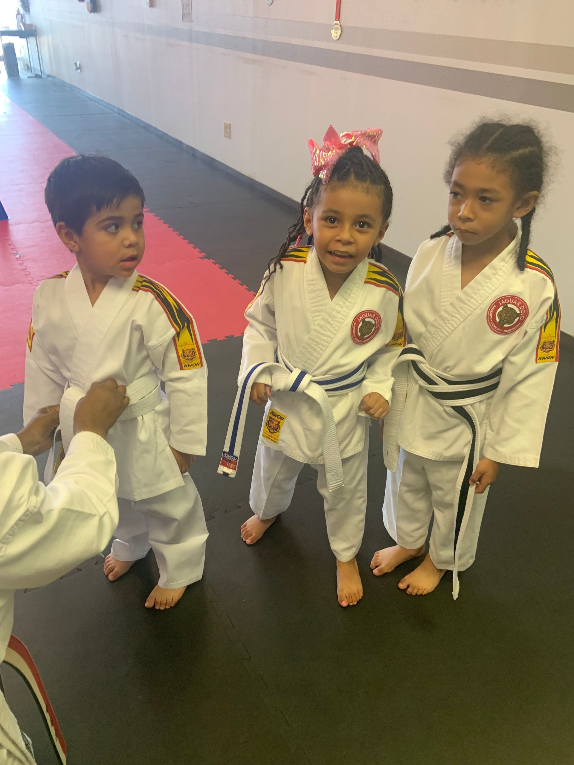 Three children in white martial arts uniforms stand barefoot in a dojo hallway.