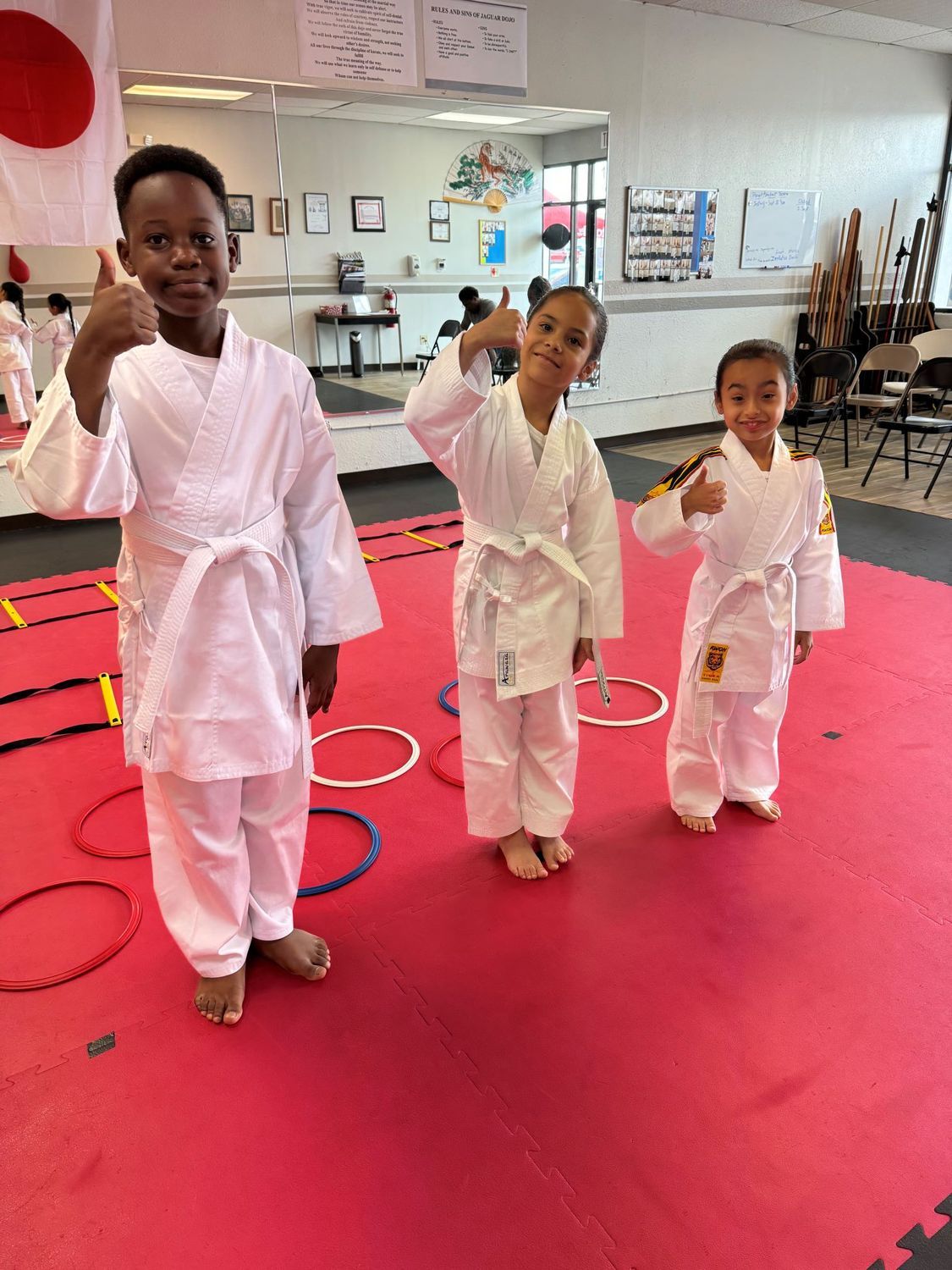 Three children in white martial arts uniforms standing on a red mat with fists raised in a dojo
