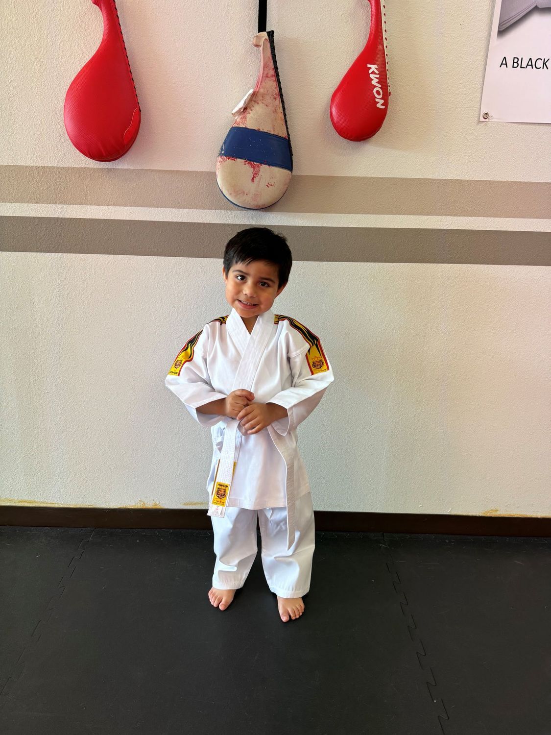 Child in a white martial arts uniform standing in a dojo beside hanging punching bags.