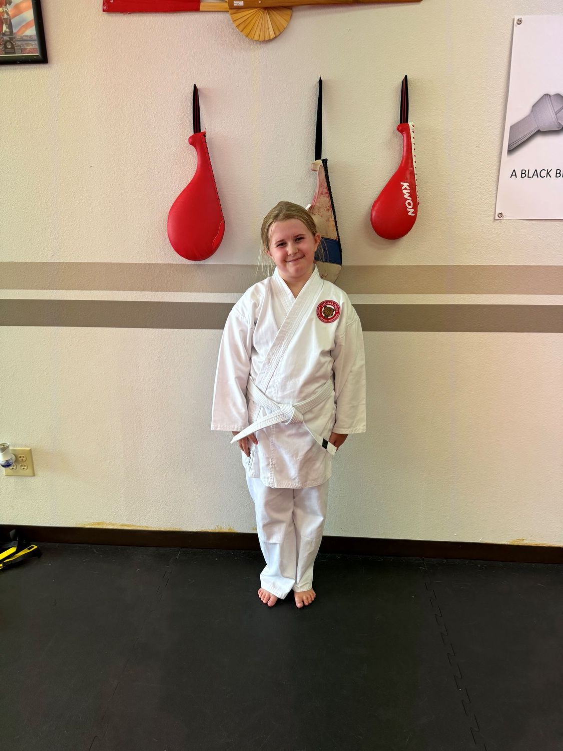 Child in a white martial arts uniform standing in a dojo with red practice paddles on the wall.