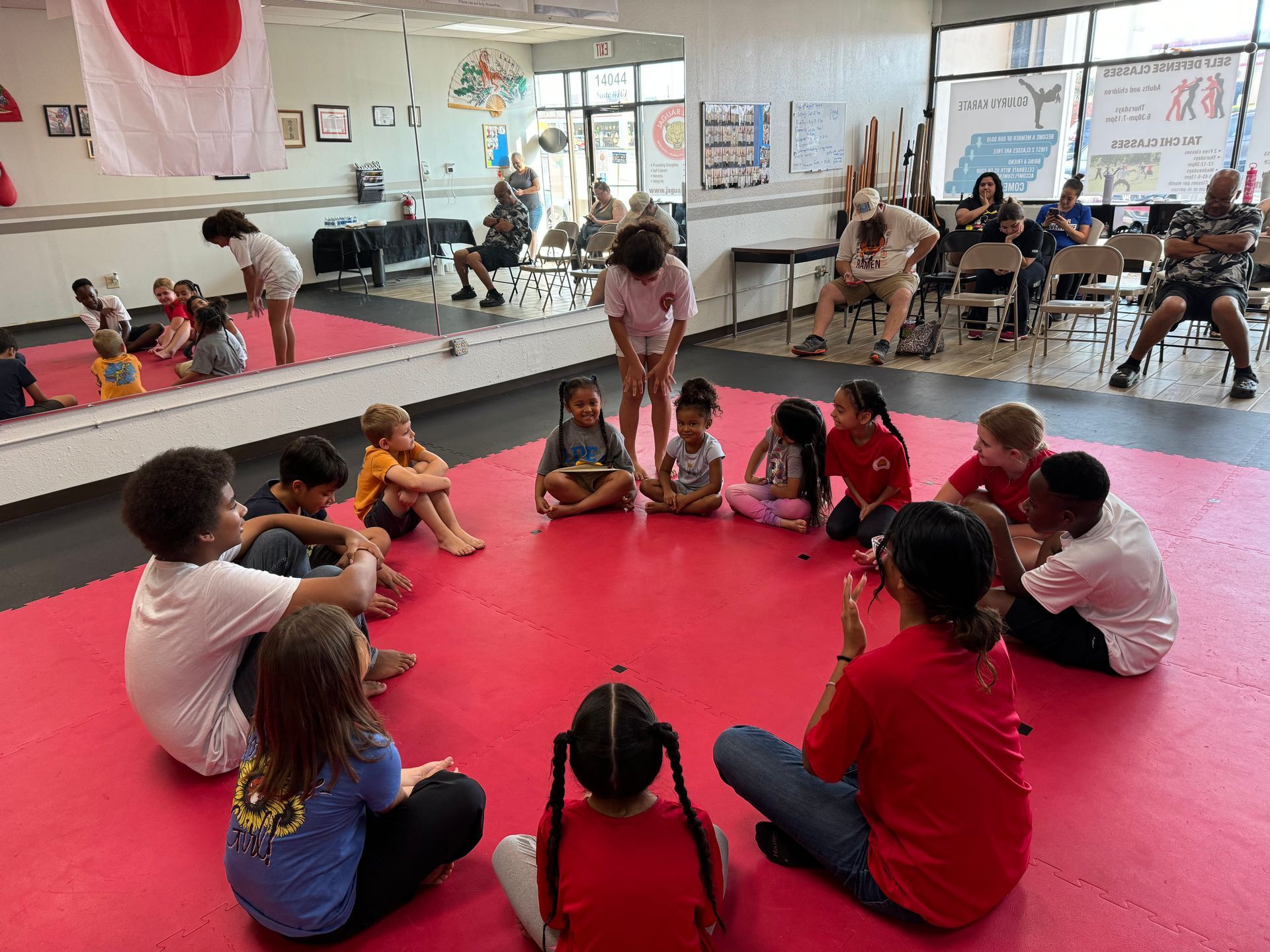Children sitting in a circle on a red mat in a dance studio, with a mirrored wall and instructor nearby
