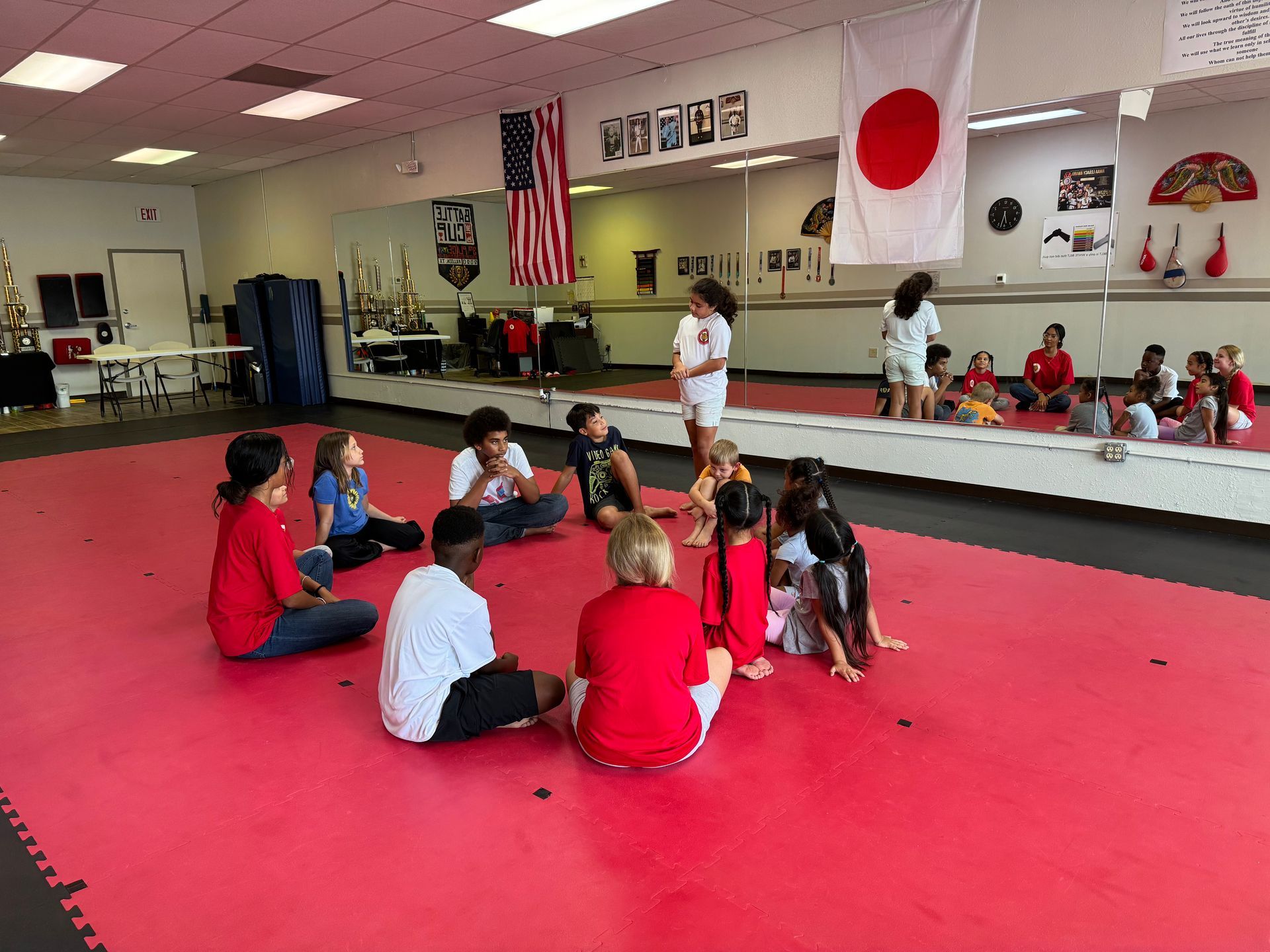 Children seated in a martial arts studio on red mats, facing mirrors and flags on the wall.
