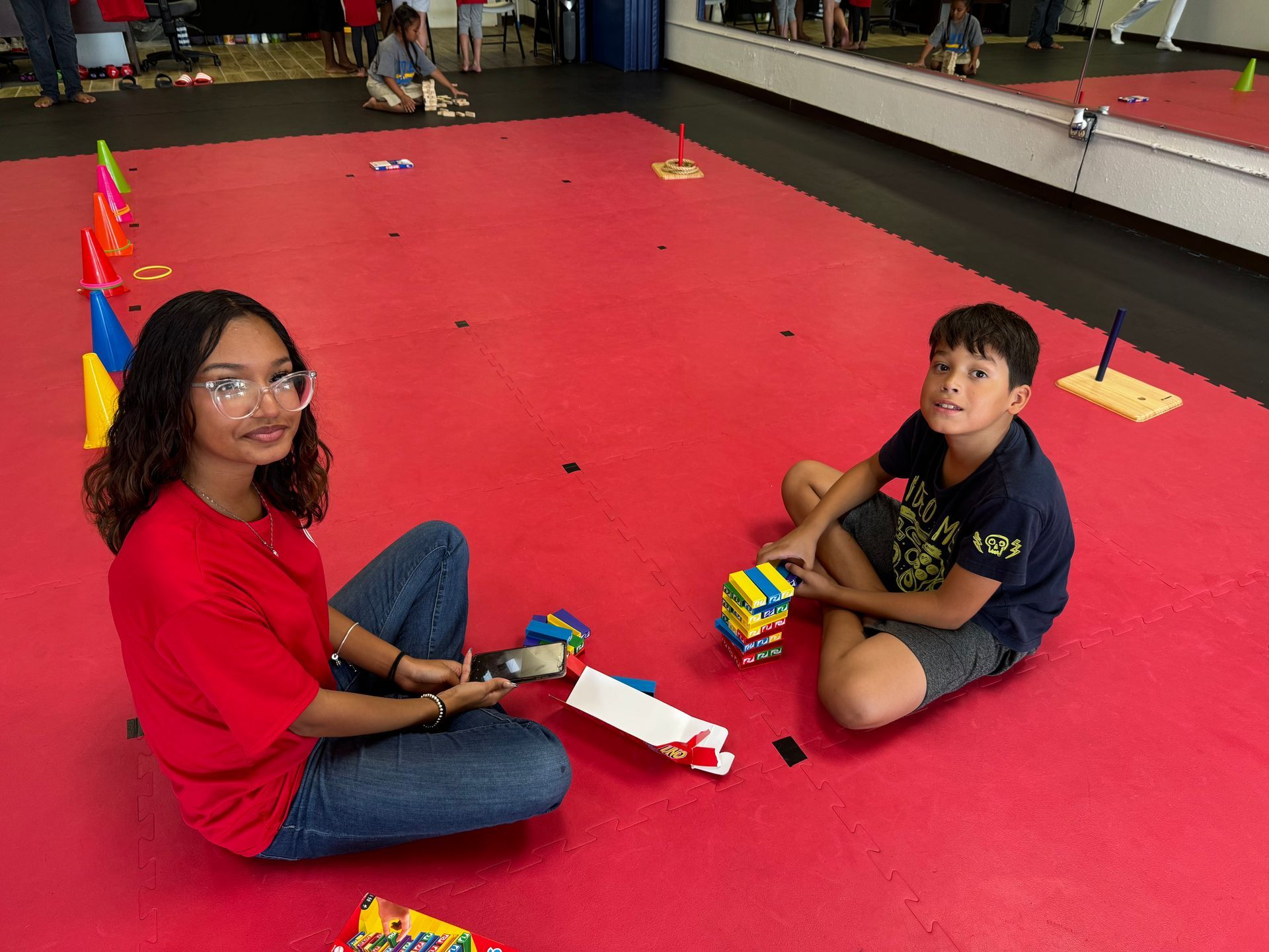 Two children sitting on a red floor, playing with colorful blocks in an indoor room.