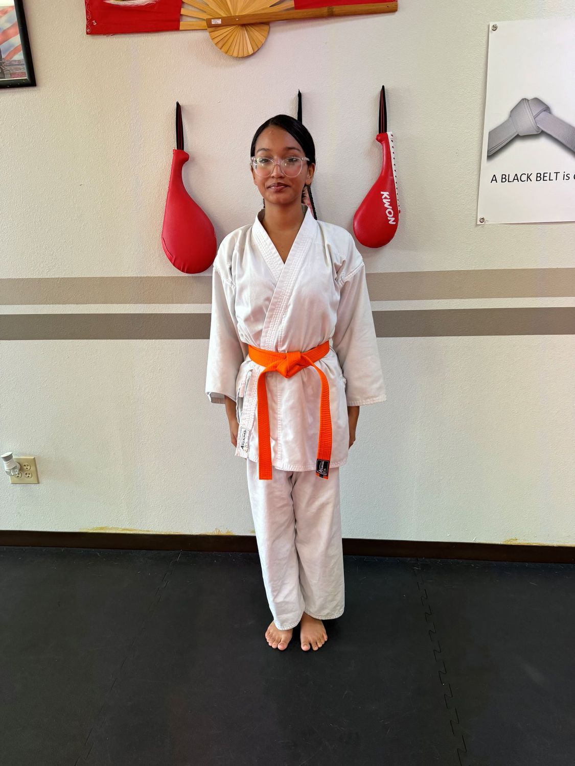 Person in a white martial arts uniform with an orange belt standing indoors against a wall.