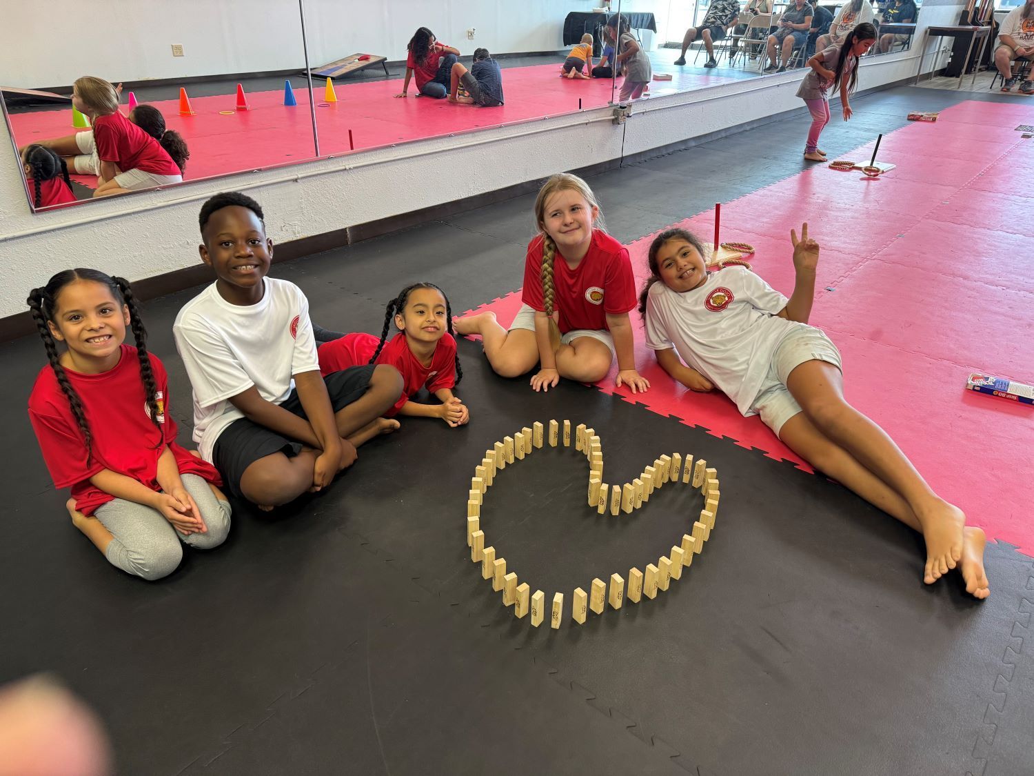 Five children sit on a gym mat beside a heart shape made of wooden blocks.