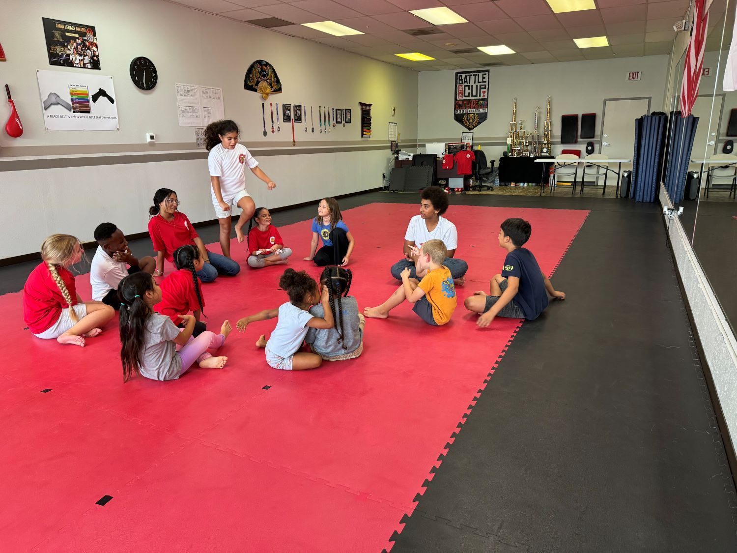 Children sitting in a martial arts studio on a red mat, listening to an instructor