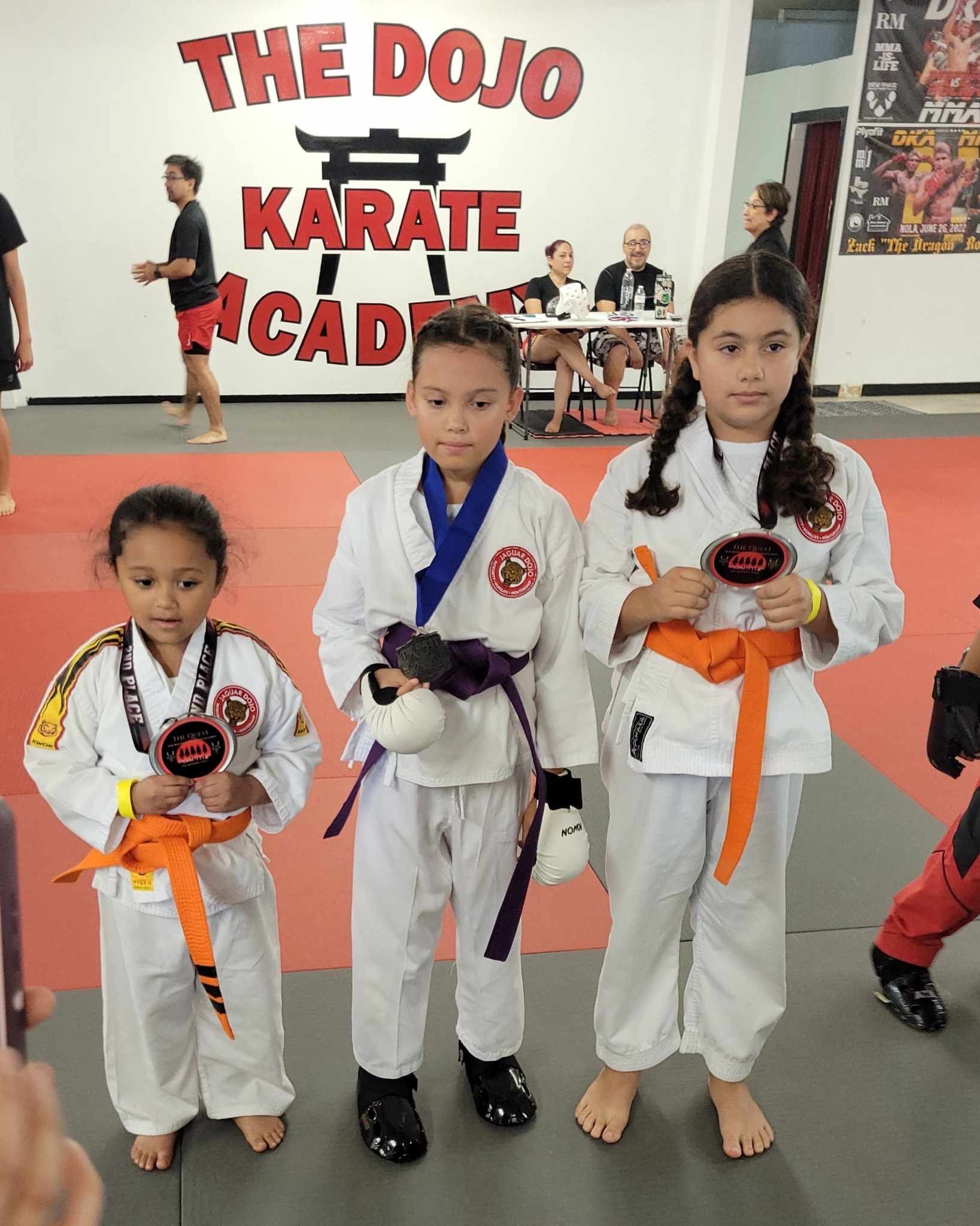 Three karate students in white gis with orange belts posing in front of The Dojo Karate Academy sign.