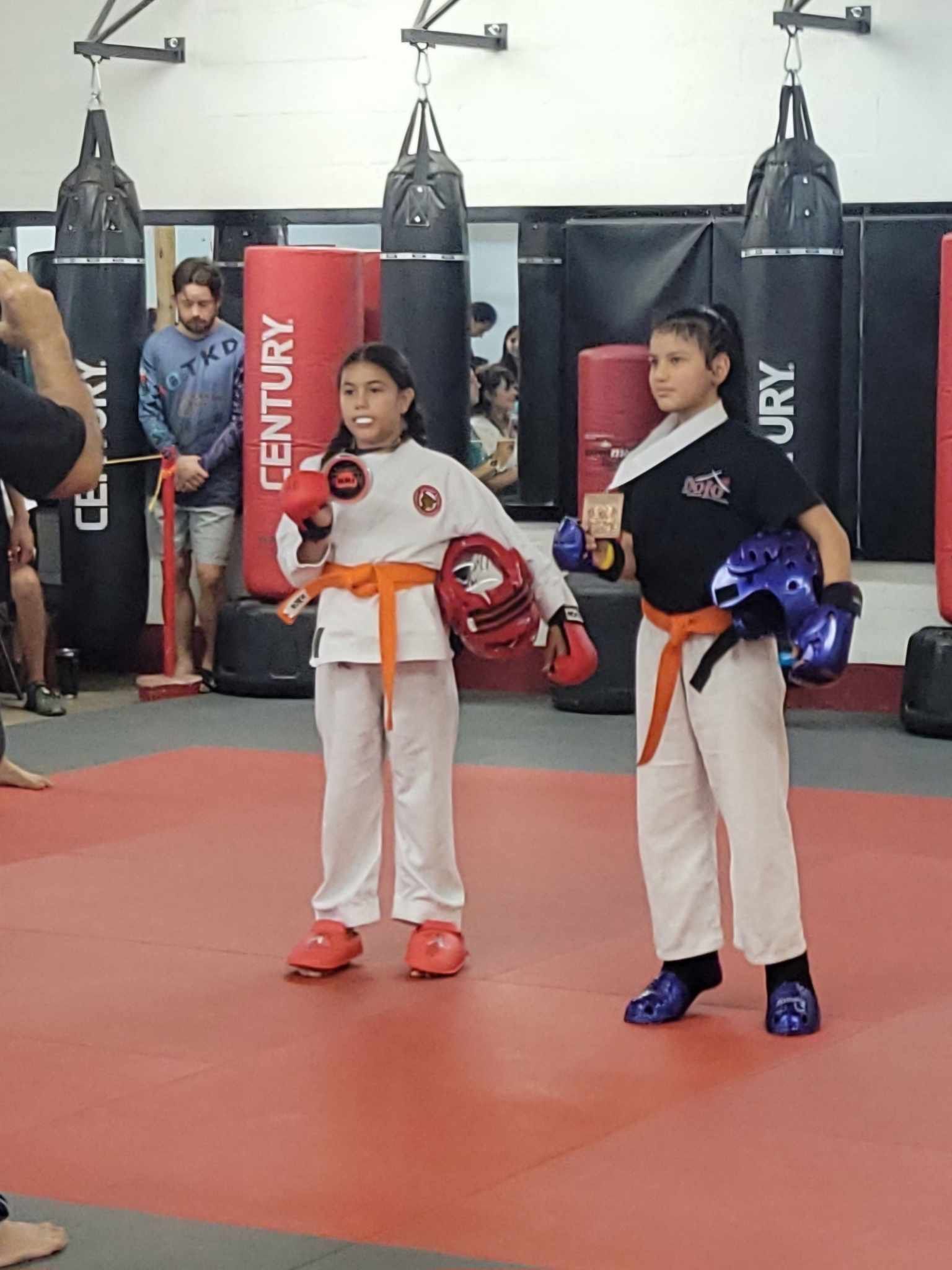 Two martial artists in white uniforms with orange belts standing in a gym with punching bags.