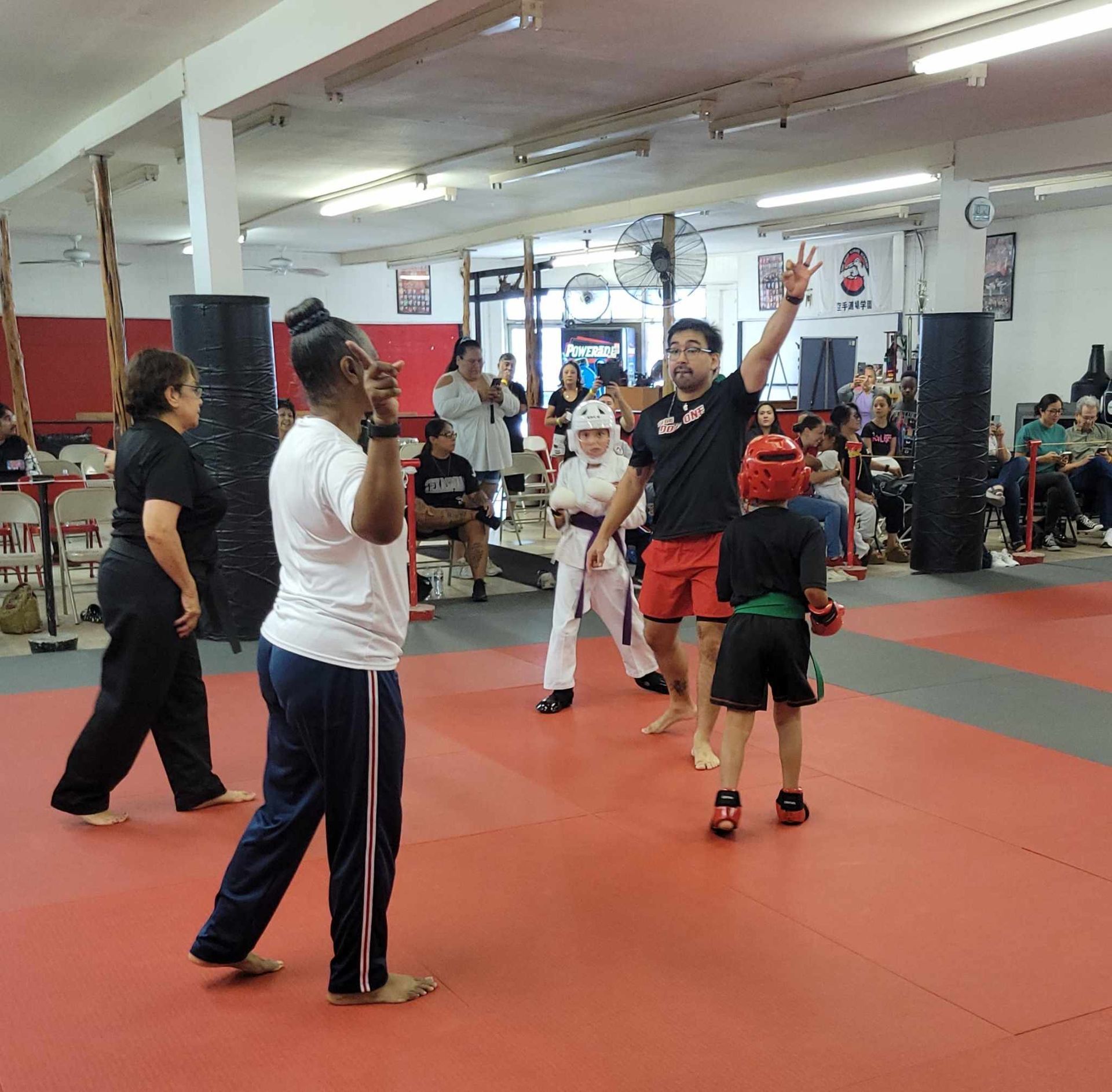 People practicing martial arts in a red-floored gym, with an instructor and students visible.